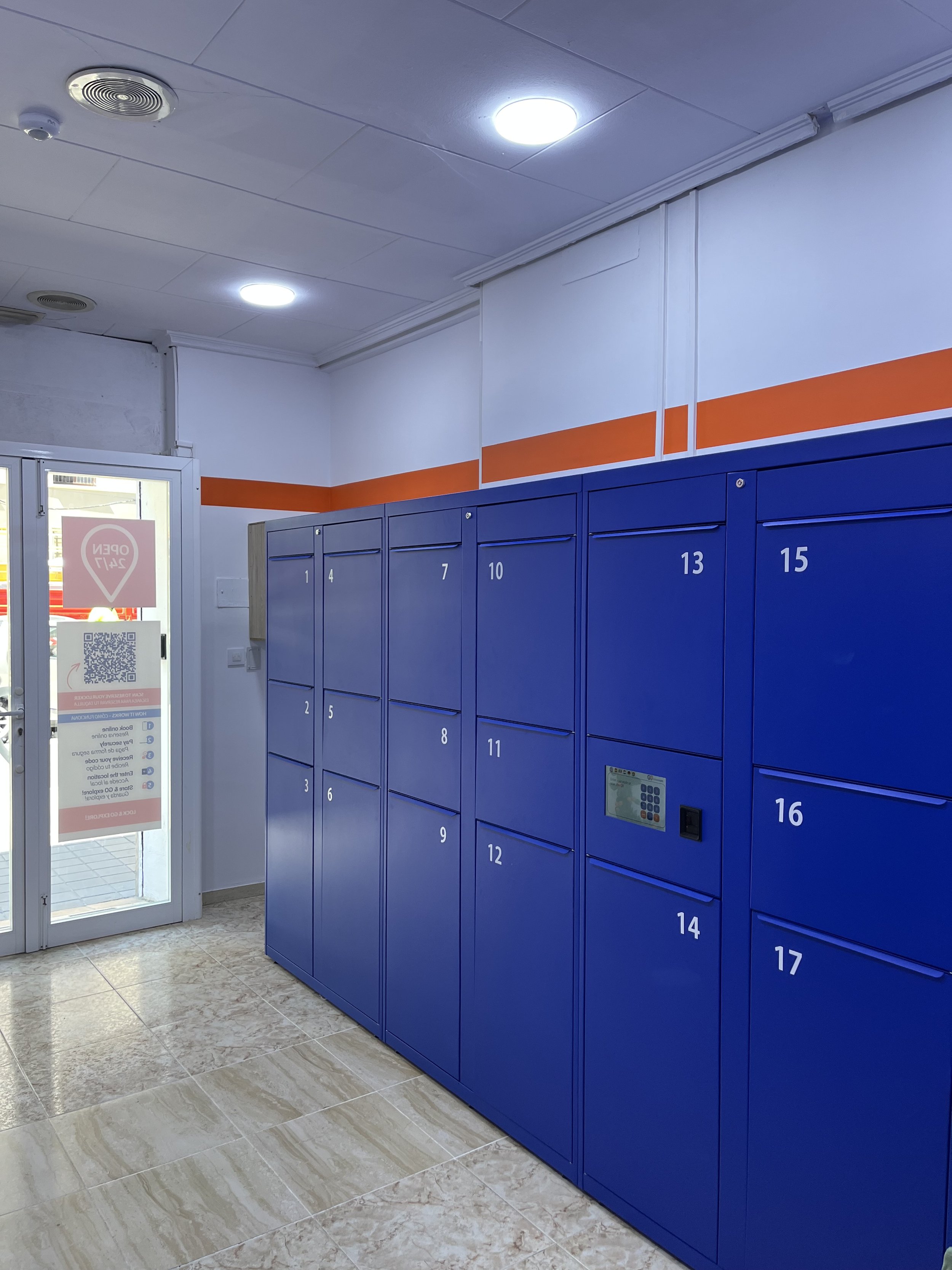 Blue storage lockers numbered 1 to 17 inside a building, with a glass door to the left that has a QR code and a sign, and a tiled floor.