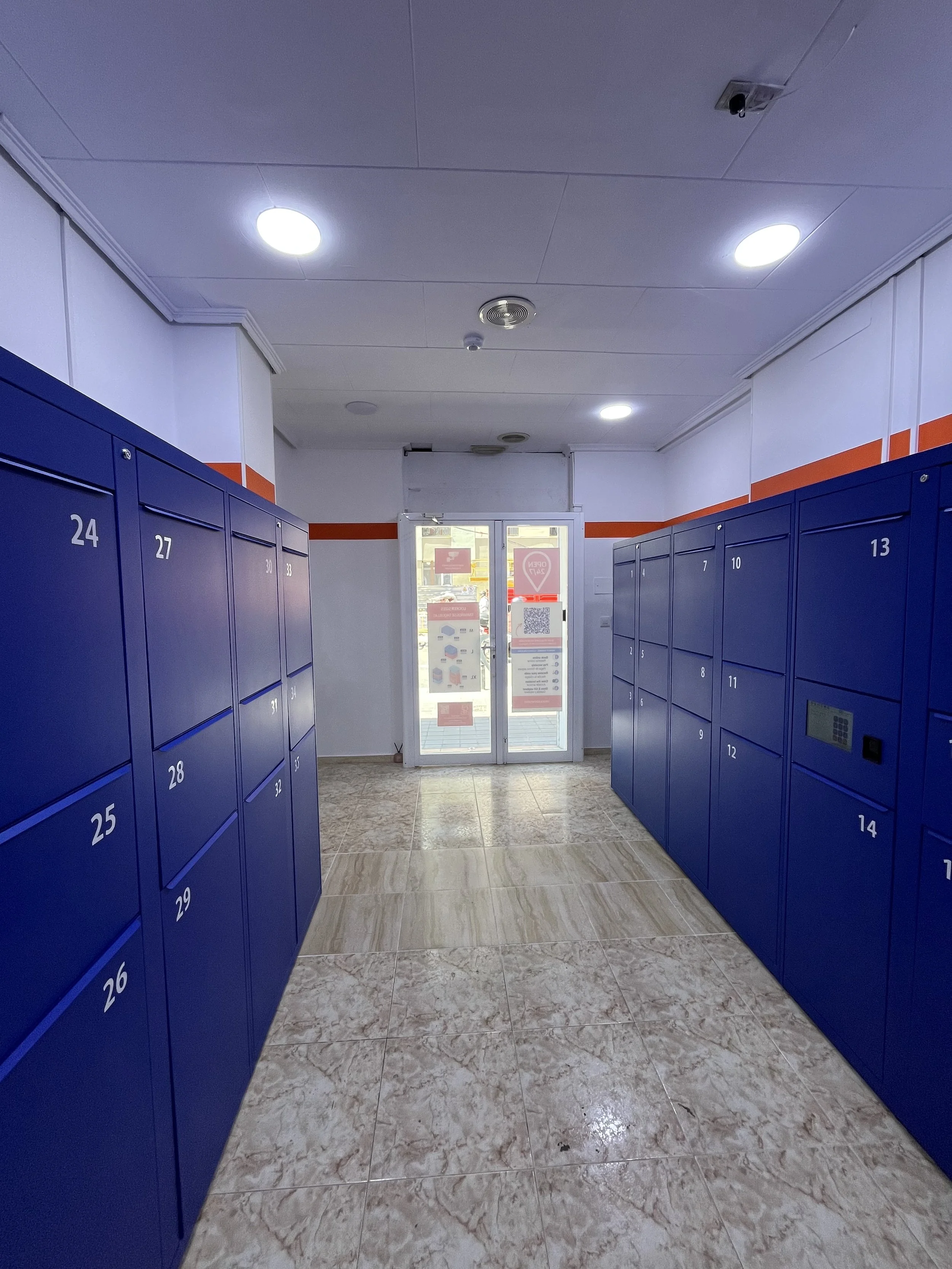 A hallway with blue lockers numbered 24 to 34 on the left and 5 to 15 on the right, leading to a glass door at the end with signage and QR code stickers, ceiling lights, and tiled floor.