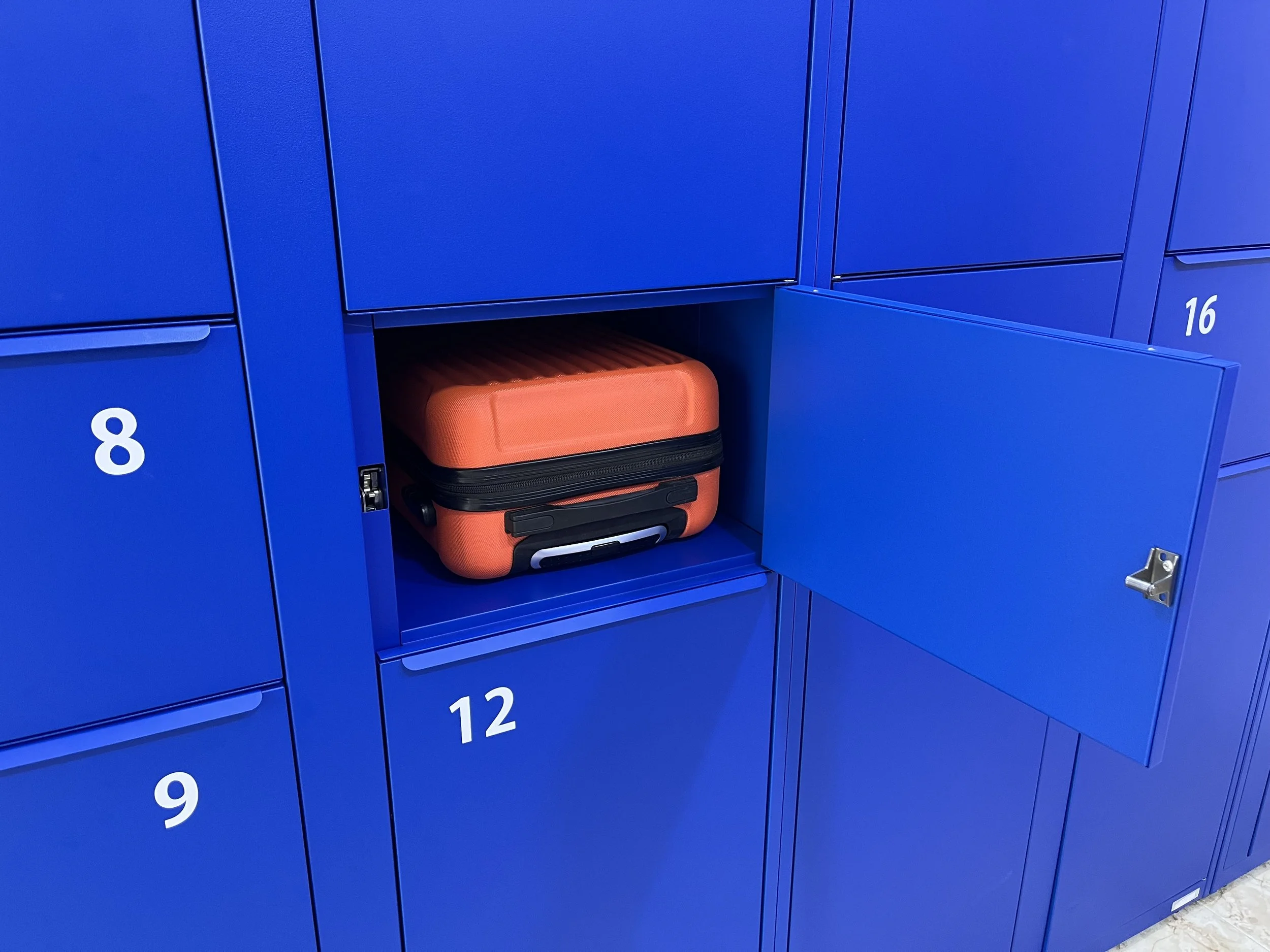 A row of blue lockers with white numbers, with locker 12 open and containing an orange suitcase.