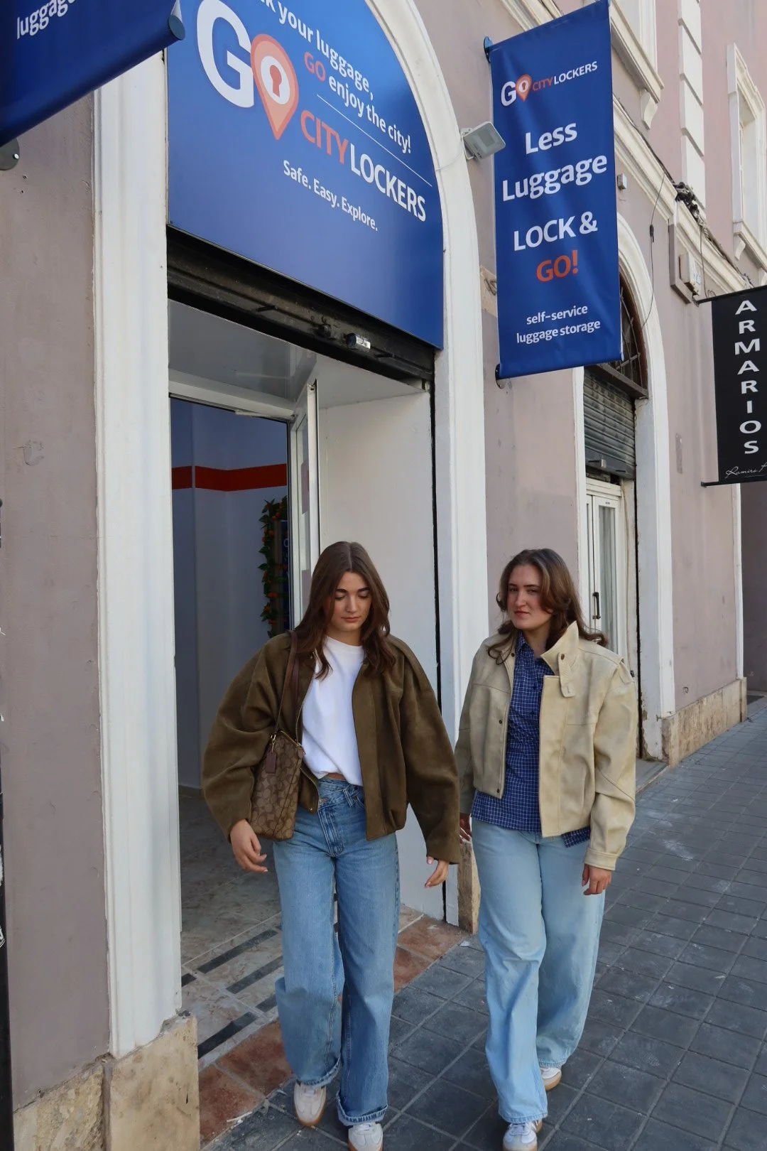 Two women walking on sidewalk in front of a luggage storage store with blue signage. One woman wears a brown jacket, white t-shirt, and jeans, carrying a patterned bag. The other wears a beige jacket, blue shirt, and jeans.