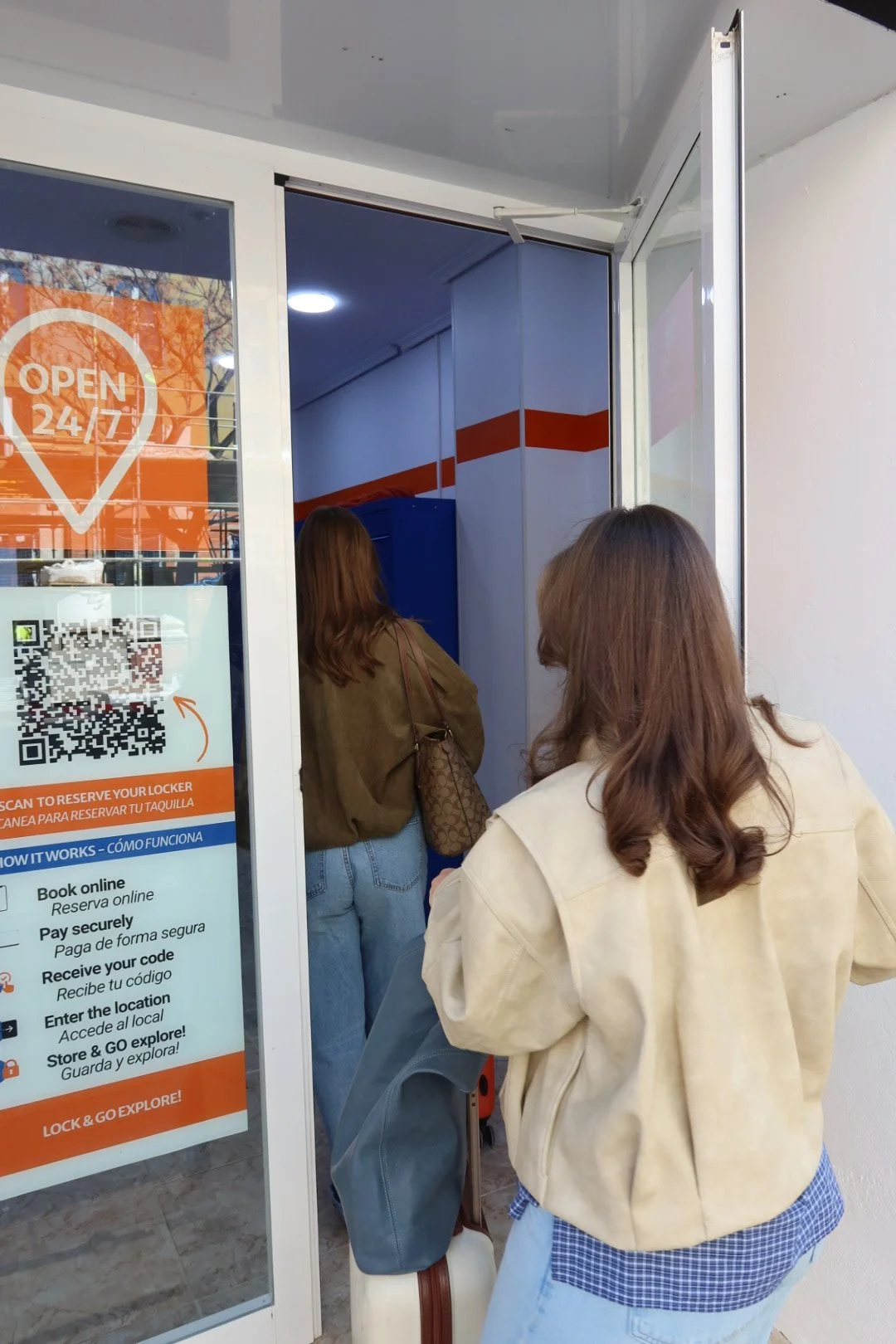 Two women standing in front of GO City Lockers, with signage on the glass door indicating 24/7 availability, including instructions on how to reserve and use lockers via QR code, and a QR code visible on the sign.