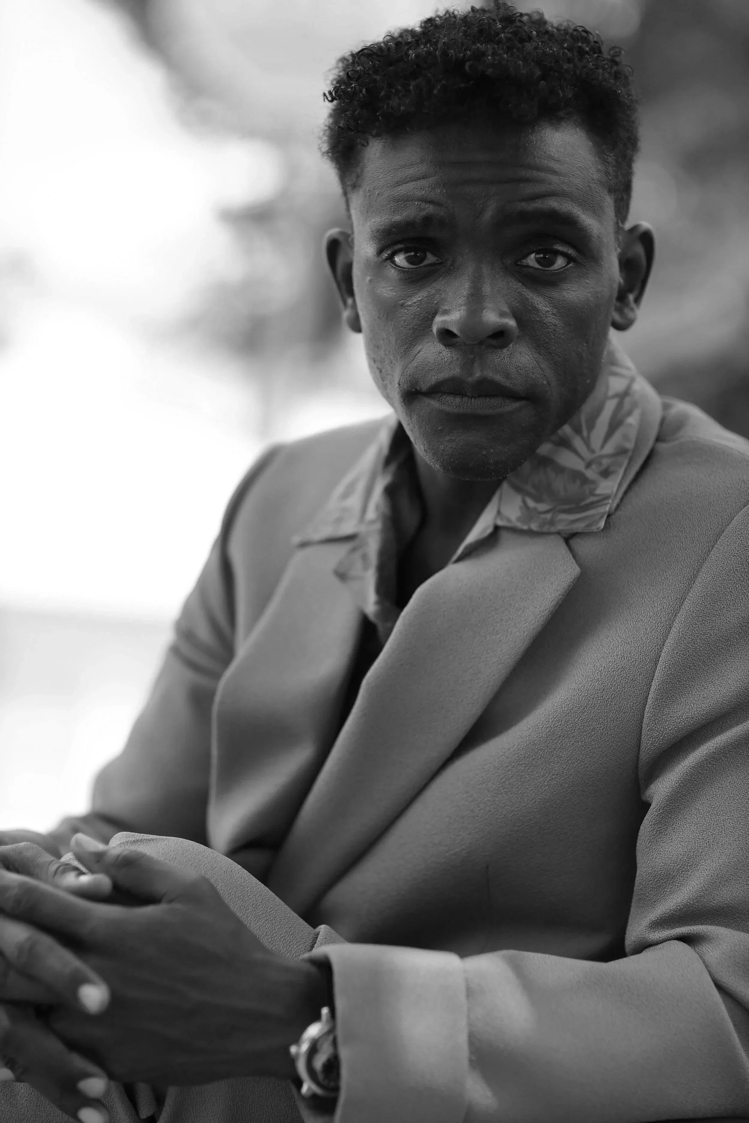 A black and white photo of a man wearing a suit and patterned shirt, looking directly at the camera with serious expression, seated with hands clasped.