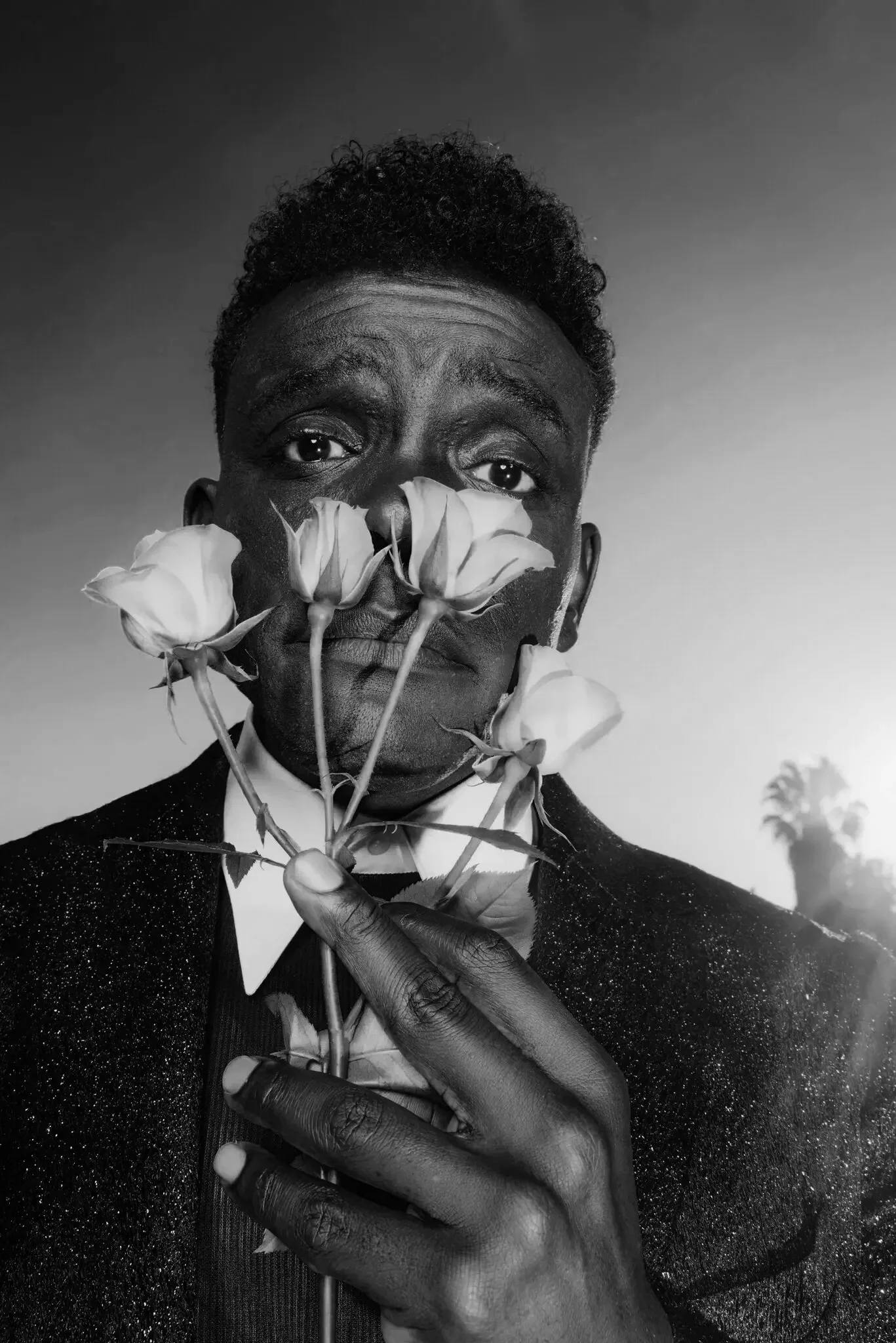 Black-and-white portrait of a man holding several roses close to his face, with some covering his mouth and nose, wearing a dark, glittery jacket and a white shirt with a bow tie. Actor. Director. Black Male.
