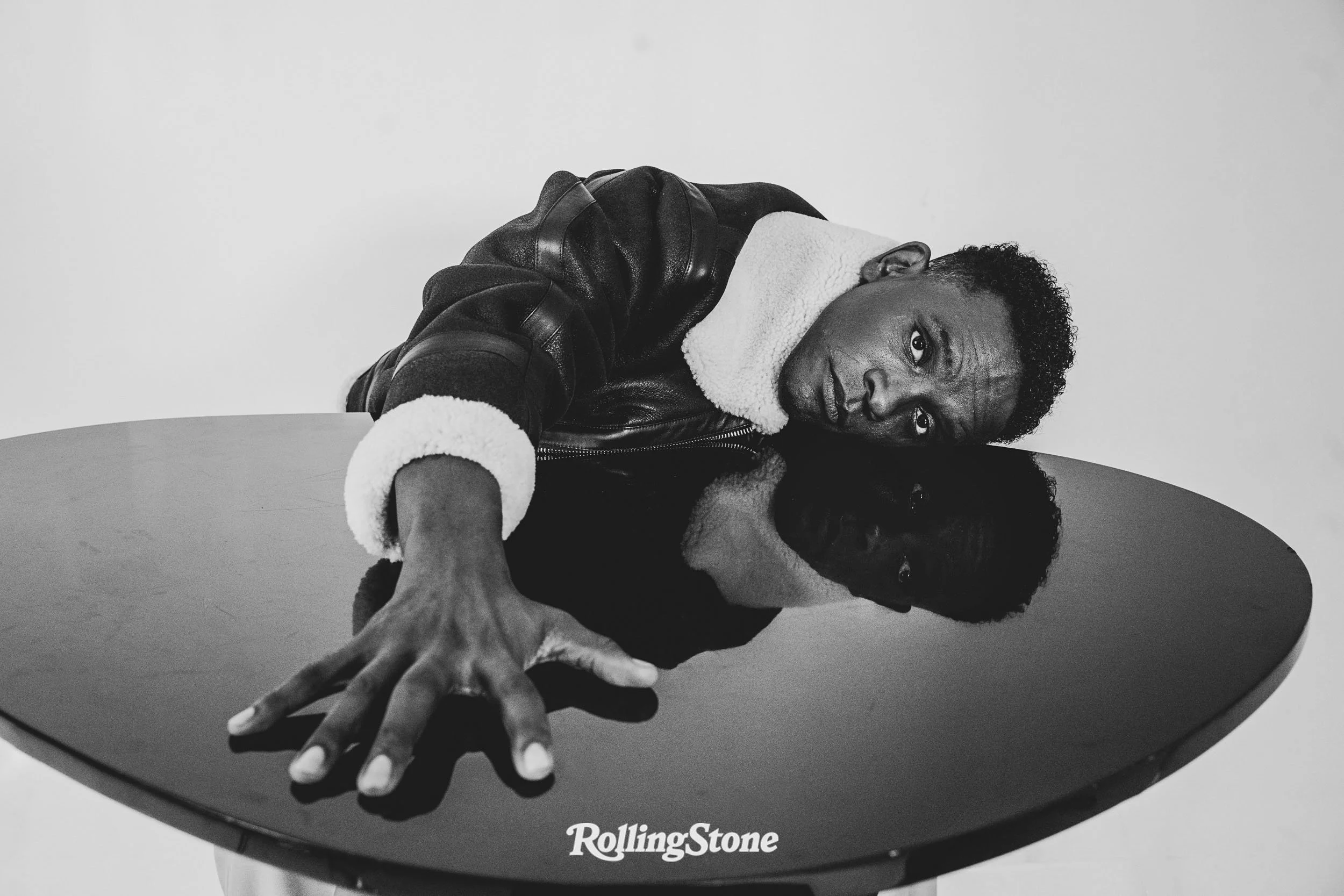 Black and white photo of a man reaching over a round table with a reflective surface, looking directly at the camera. The man is wearing a dark jacket with a shearling collar. Actor. Black Male.
