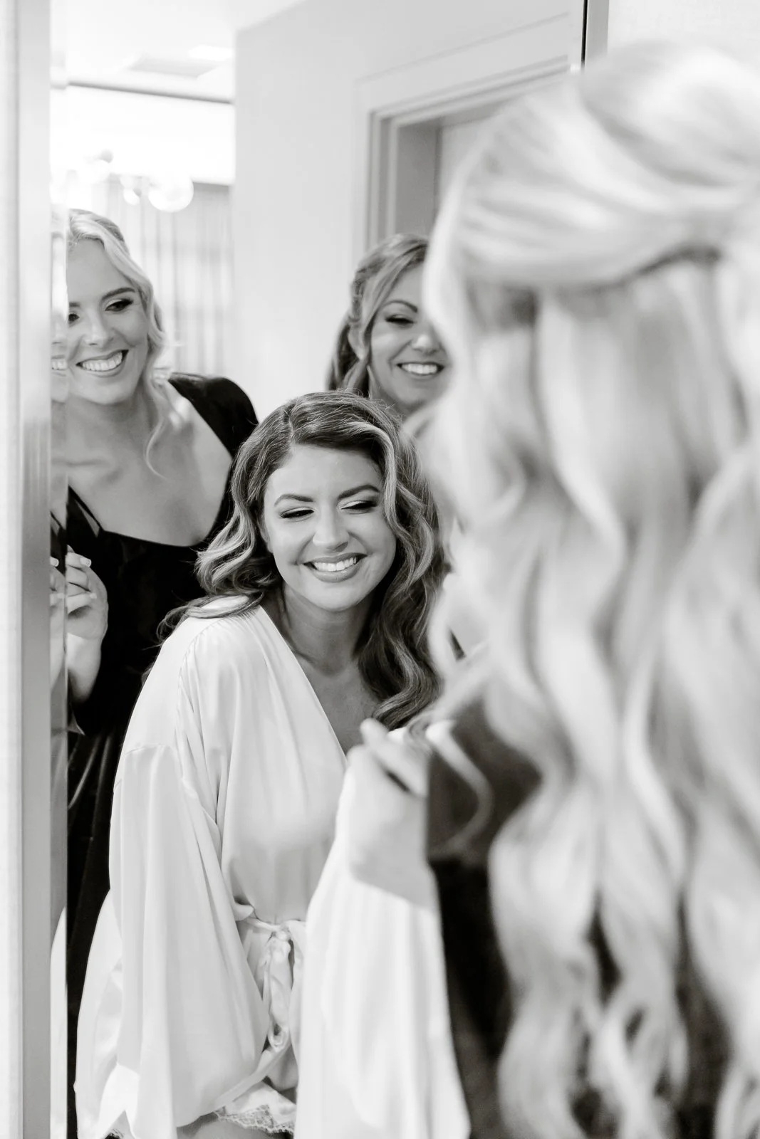 Four women looking at their reflections in a mirror; they are smiling and appear happy. One woman in the foreground has wavy hair and is wearing a white robe. The other women are behind her, also smiling.