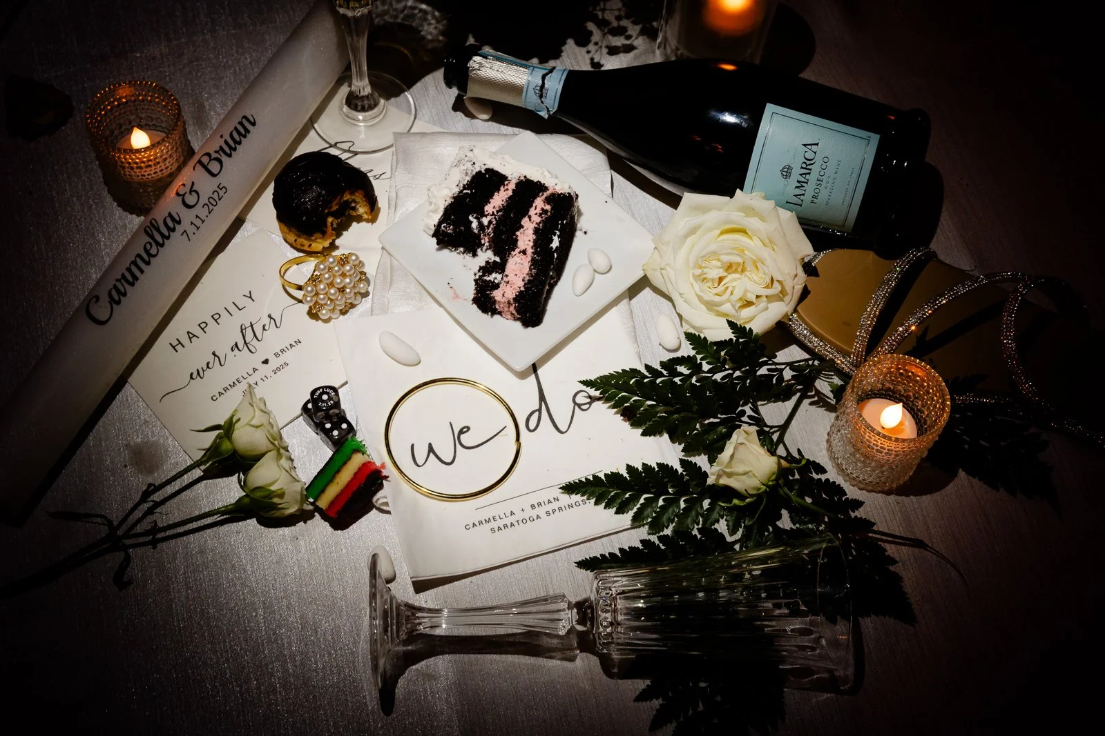 Wedding table display with a name plate for Carmella and Brian, a black and pink layered cake slice, white roses, candles, champagne bottle, glasses, a white 'we do' sign, fern leaves, a small Italian flag, and wedding details including date and loca