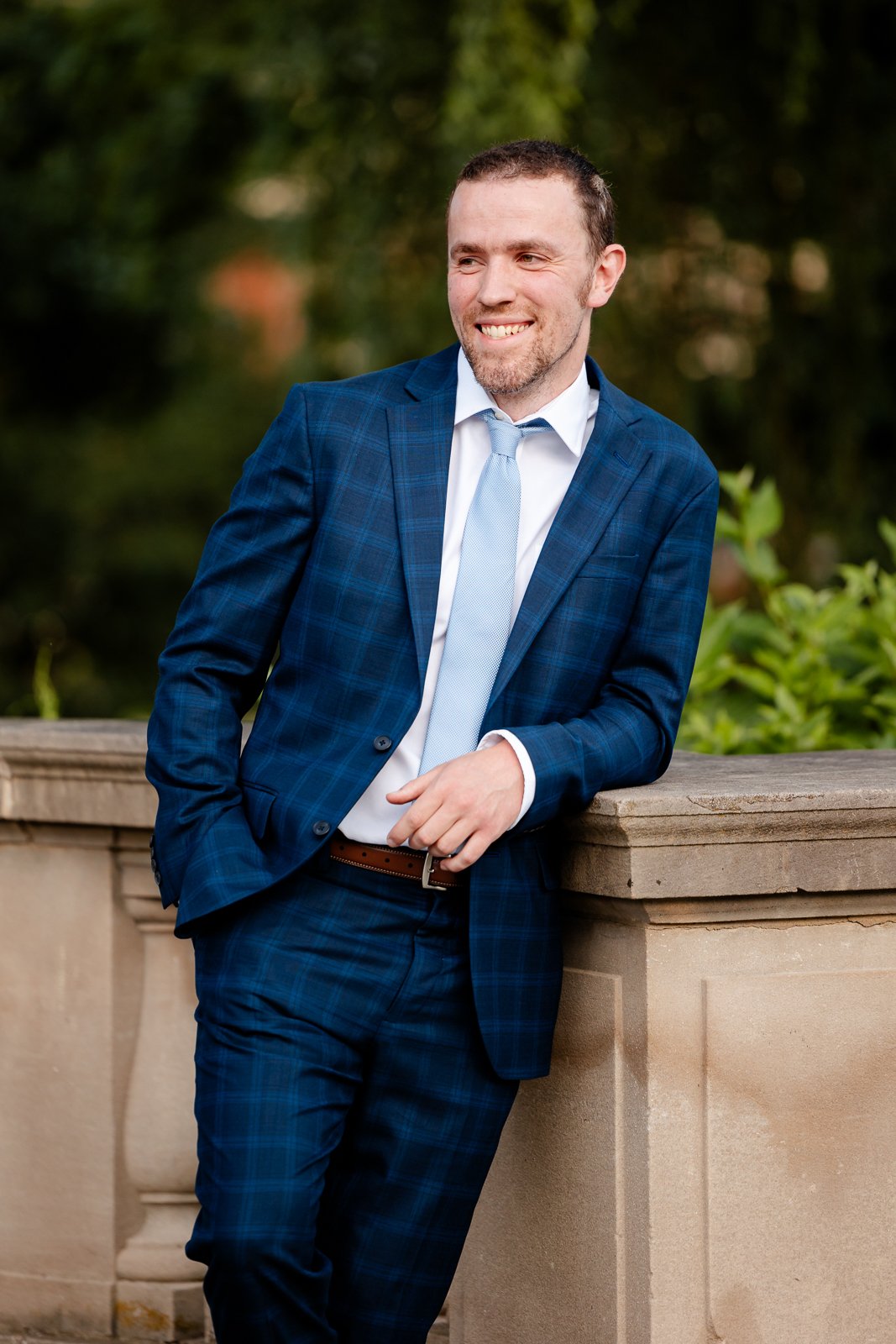 A man dressed in a blue plaid suit with a light blue tie, leaning on a stone railing outdoors, smiling, with blurred greenery in the background.
