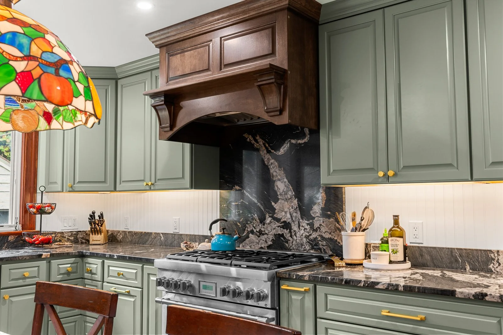Kitchen with green cabinets, black marble backsplash, and a stainless steel oven. A blue kettle is on the stove, and a stained glass lamp is hanging from the ceiling. Various kitchen utensils and condiments are on the countertop.