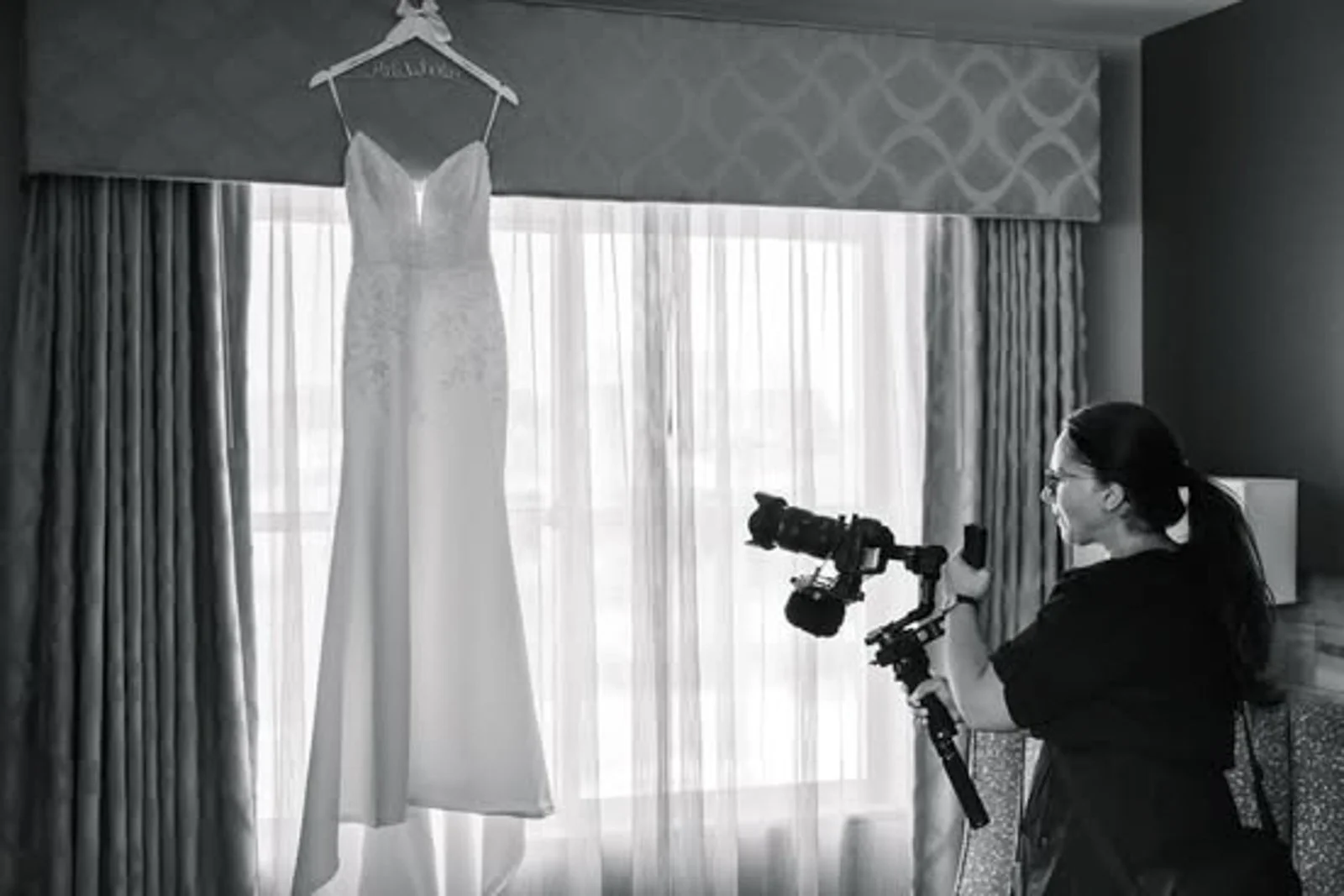 A woman is taking photos of a white wedding dress hanging from a hanger in front of a window with curtains.
