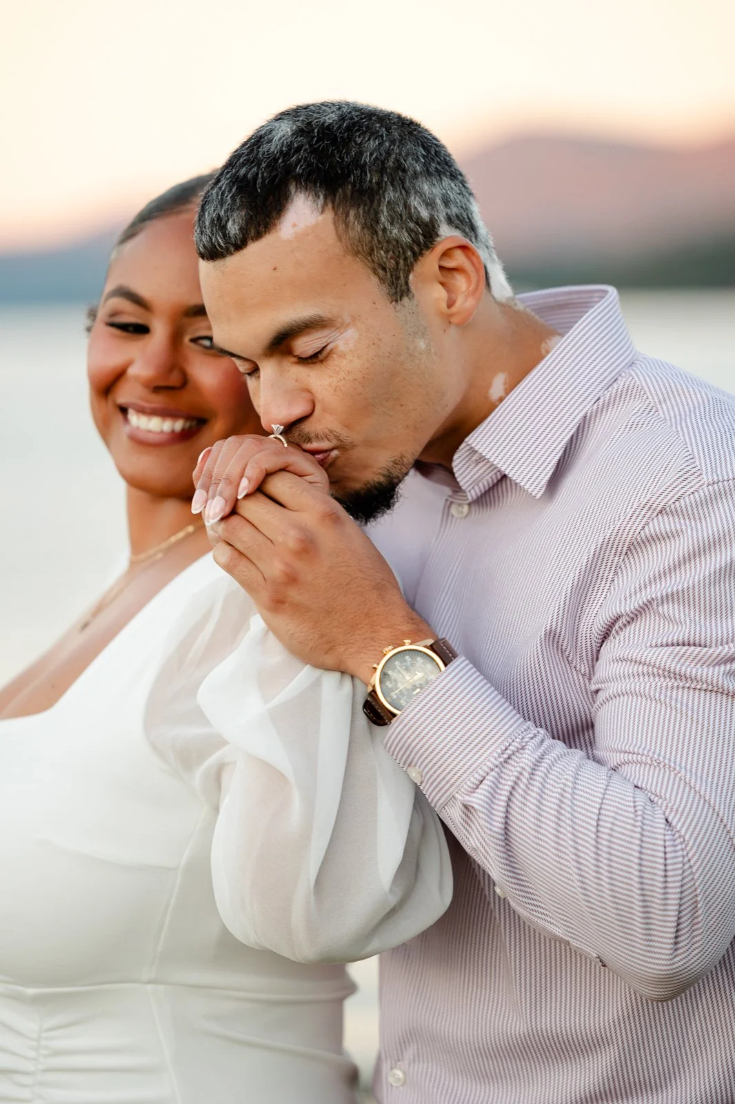 A man kissing a woman's hand, both smiling, outdoors near a body of water at sunset.