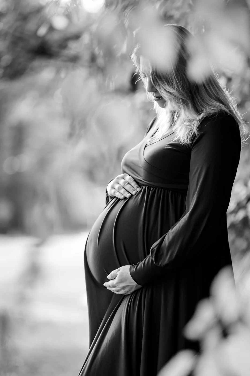 A pregnant woman in a long dress gently cradling her belly with both hands outdoors, with blurred foliage surrounding her.