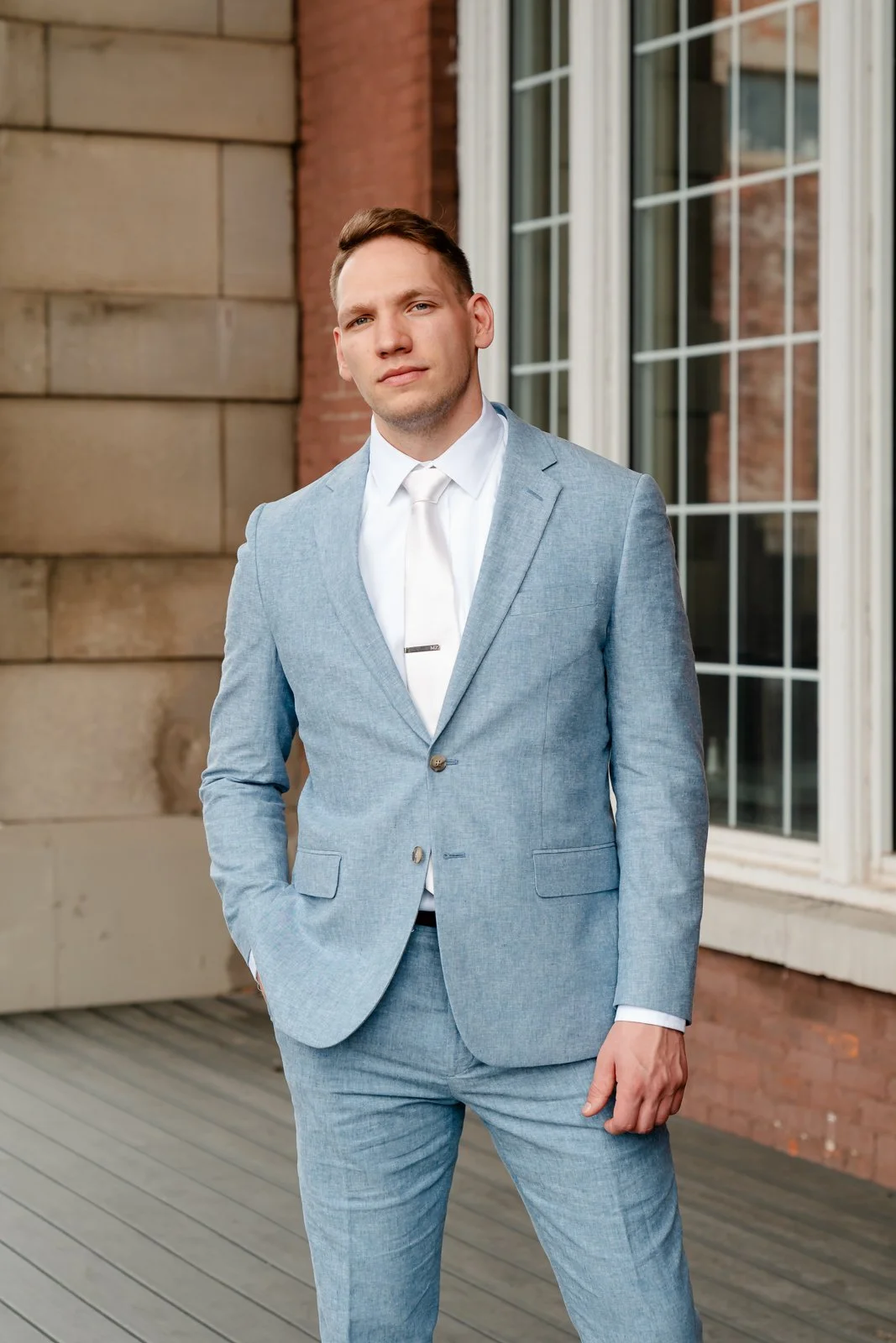 A man in a light blue suit, white shirt, and matching tie stands outdoors on a wooden deck in front of a brick building with large window panes.