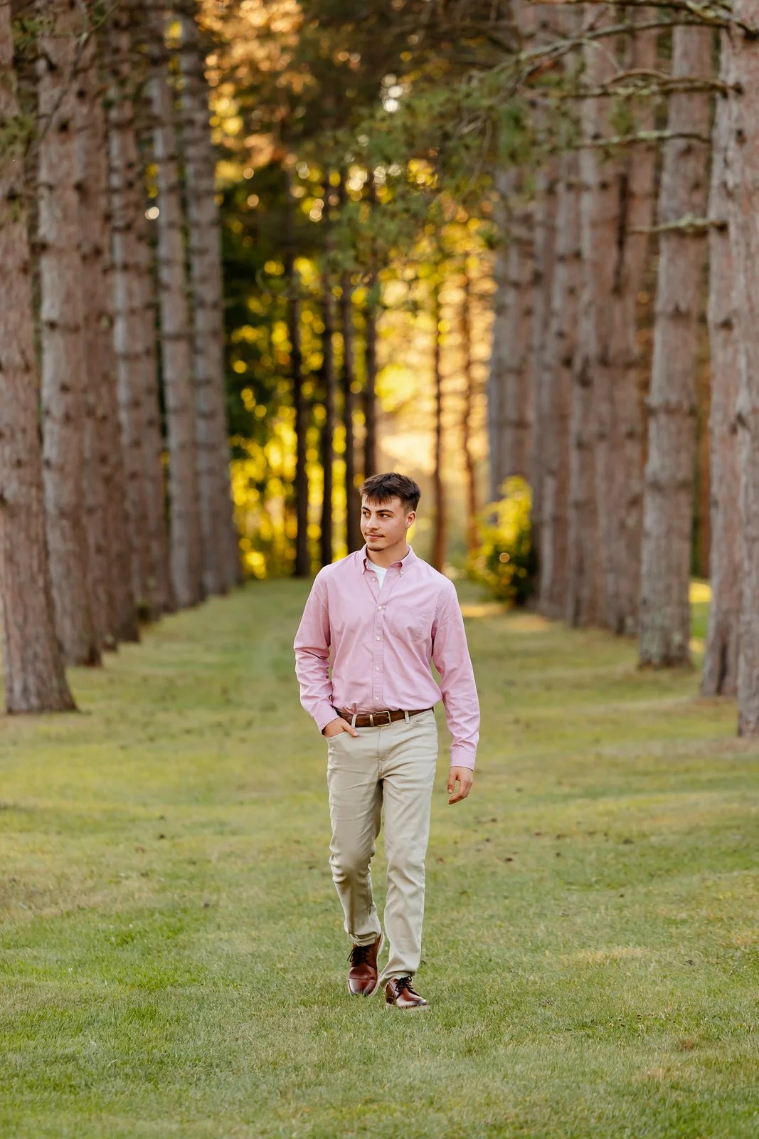 A young man walking along a grassy path lined with tall trees in a forest during sunset.