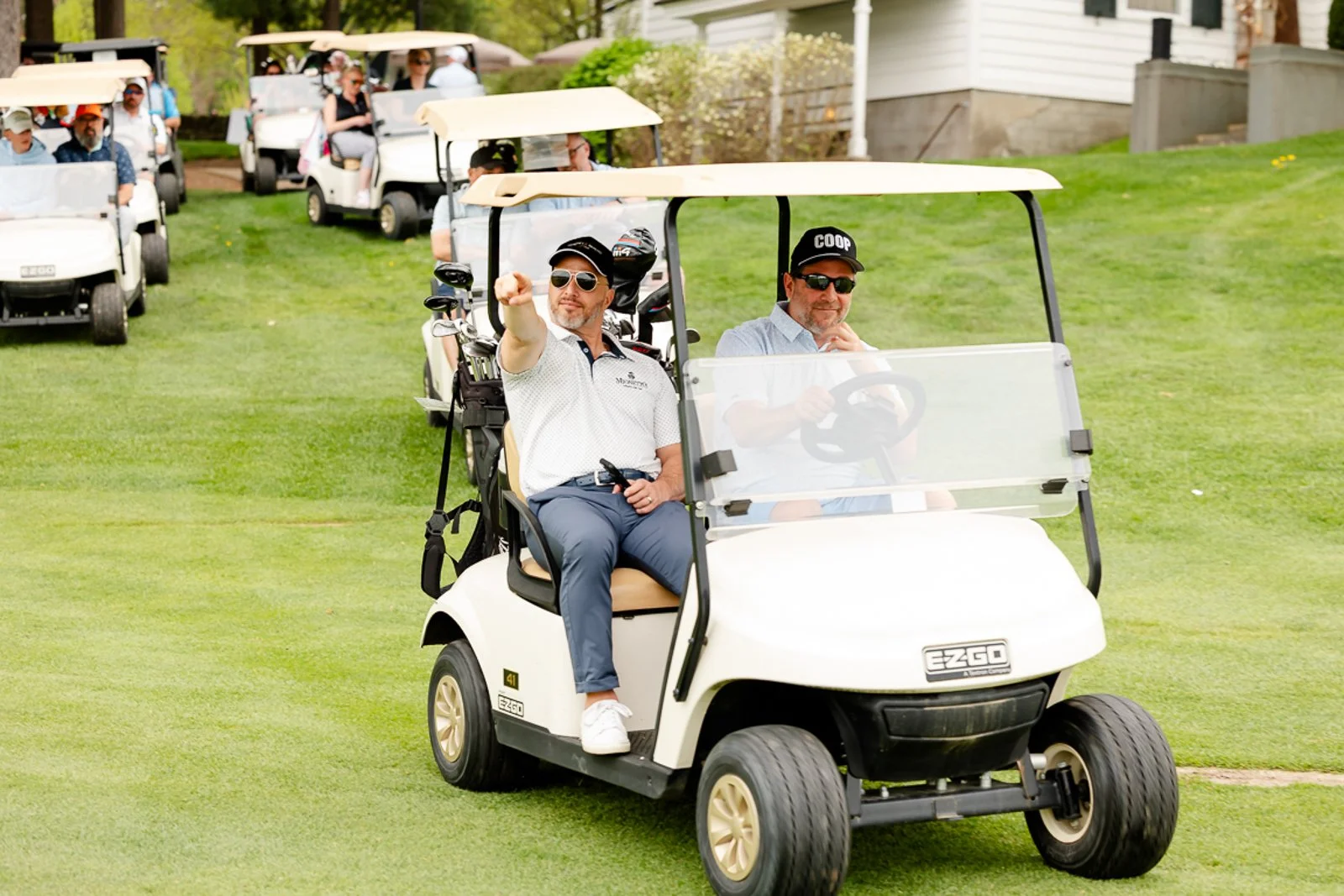 Men riding in golf carts on a golf course, with others following behind.