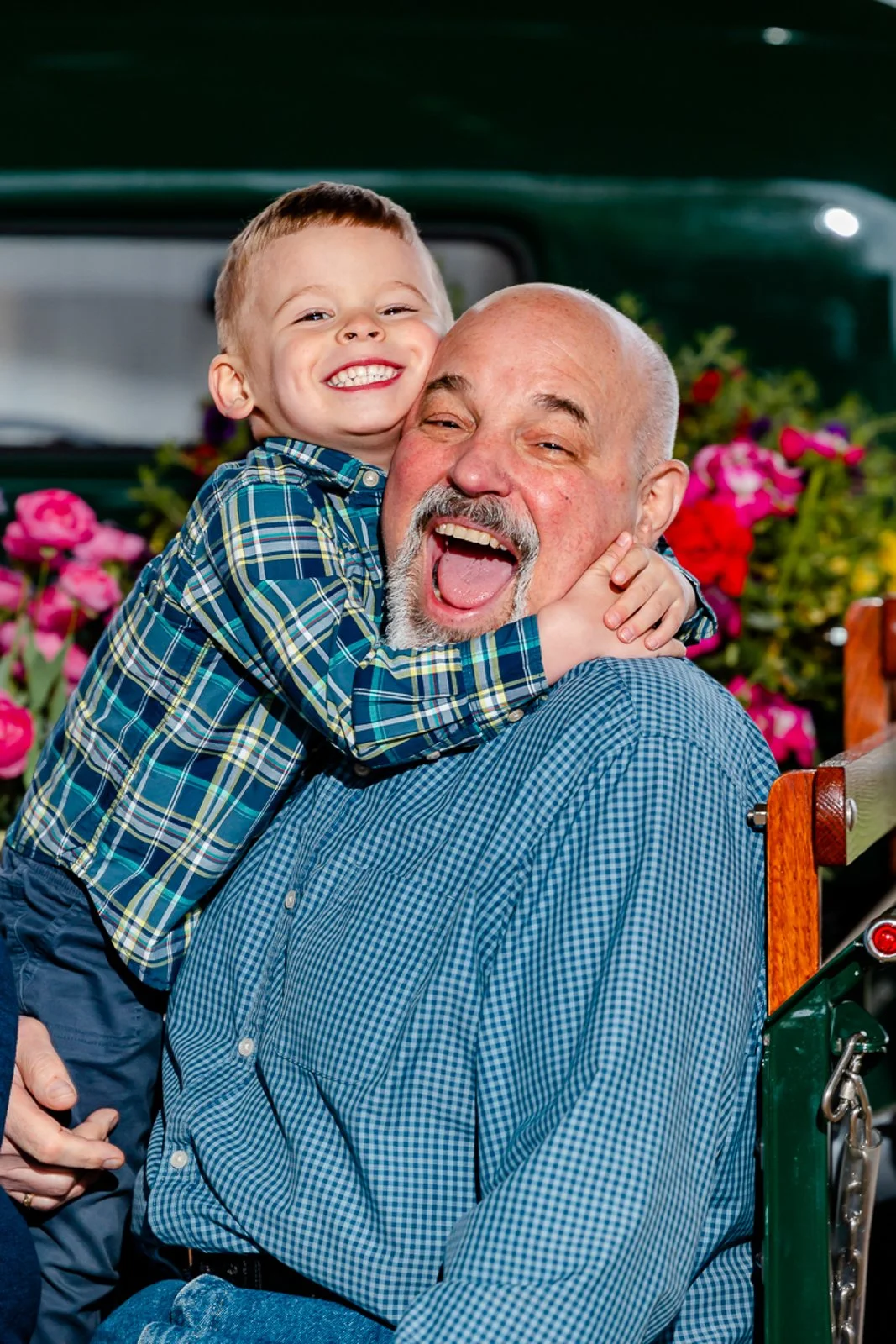 A young boy hugging an older man in a wheelchair, both smiling joyfully, with colorful flowers in the background.