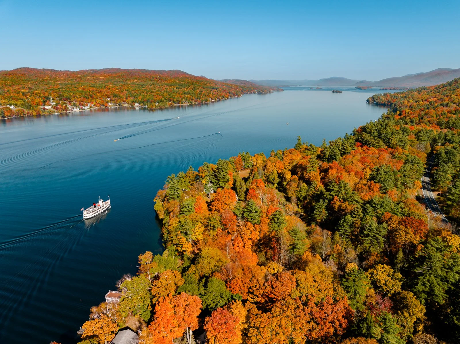 Aerial view of a large lake surrounded by colorful autumn trees and hills, with boats sailing on the water.