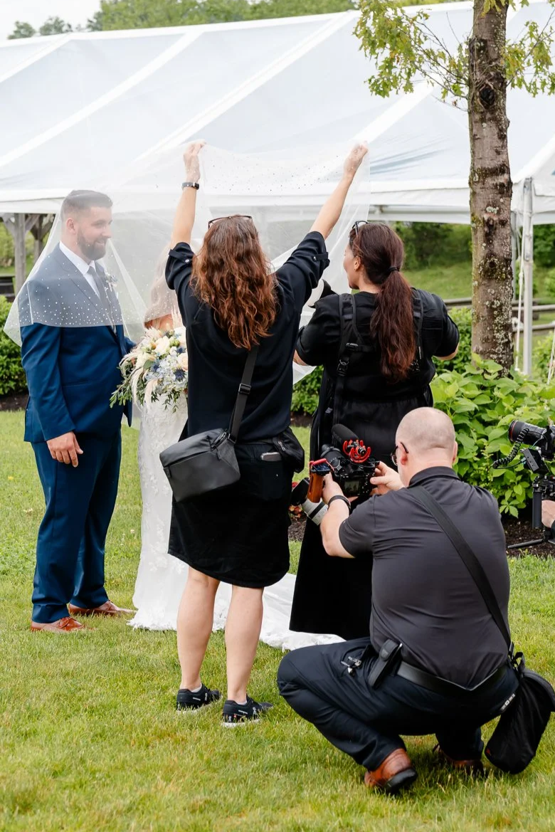 Photographers preparing to take a wedding photo of a groom in a blue suit and a bride holding a bouquet, under an outdoor tent.