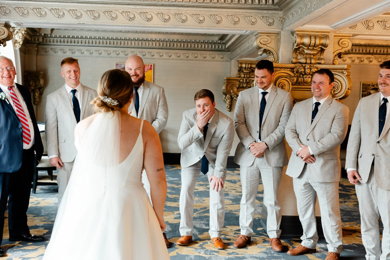 Bride with a white dress and updo hairstyle is facing a group of seven men in suits, some are smiling while one covering his mouth, in an ornate indoor setting.