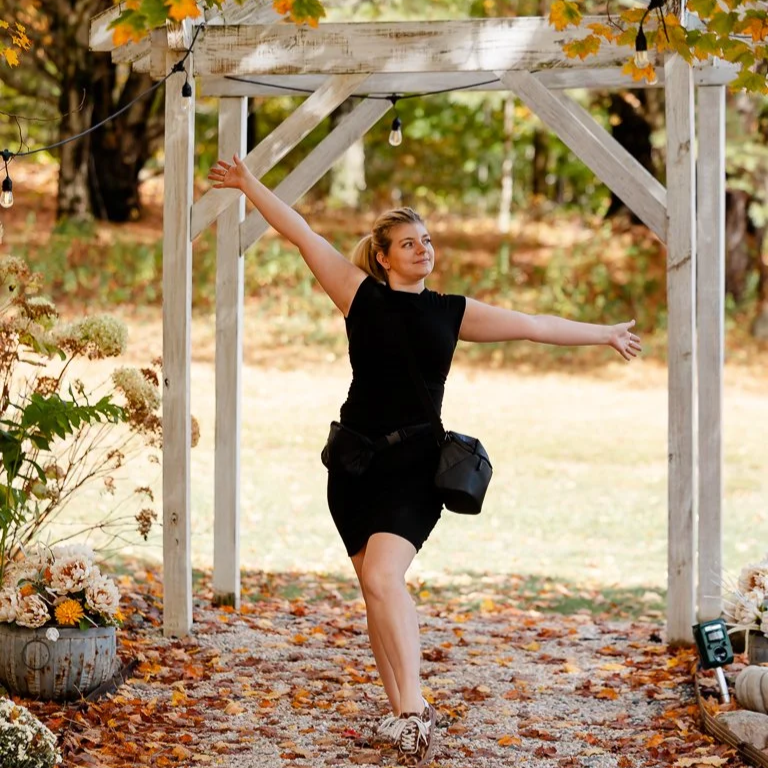 A woman in a black dress and sneakers performs a dance move outdoors under a wooden pergola decorated with string lights and autumn leaves.