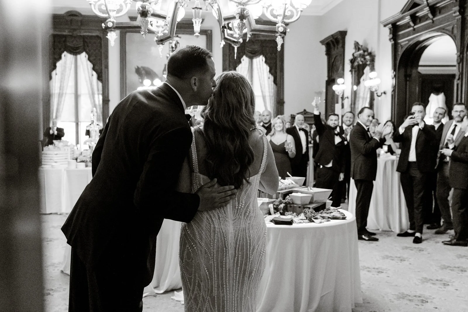 A couple, possibly at a wedding reception, stands close together with the man whispering into the woman's ear in front of a decorated table. Guests in formal attire are clapping and taking photos in a grand, elegant room with ornate woodwork, large windows, and a chandelier.