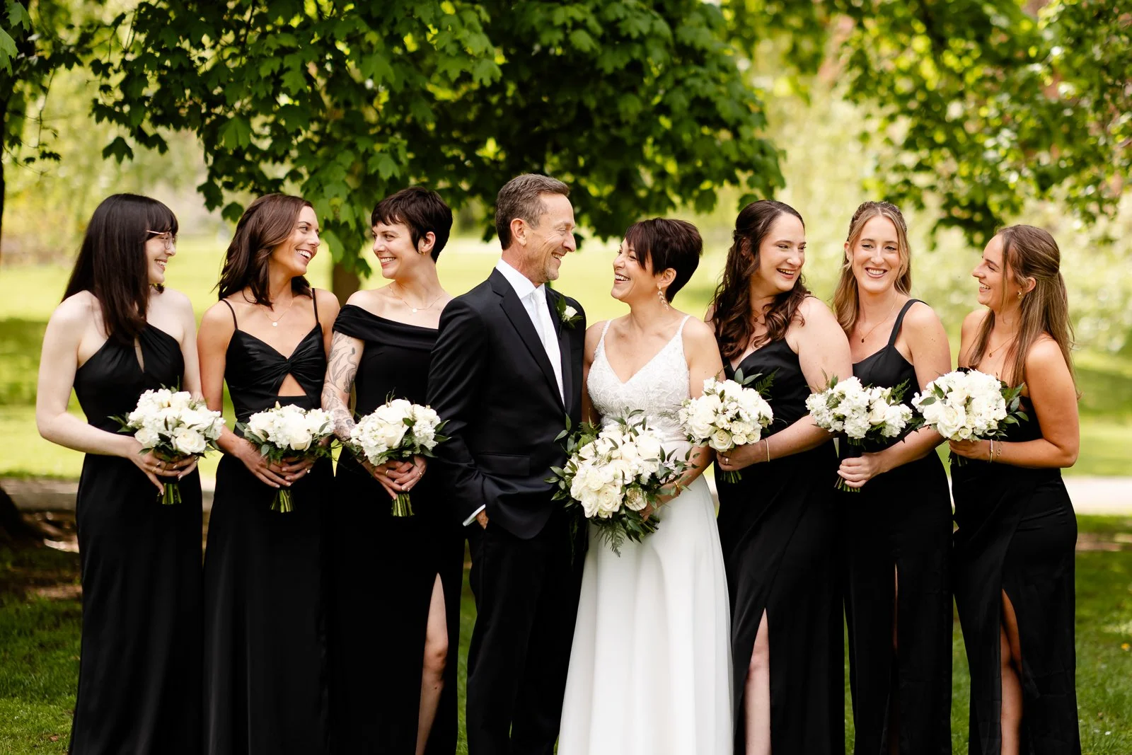A wedding party outdoors with a bride and groom in the center, surrounded by six women in black dresses holding white bouquets, all smiling and looking at each other under green trees.