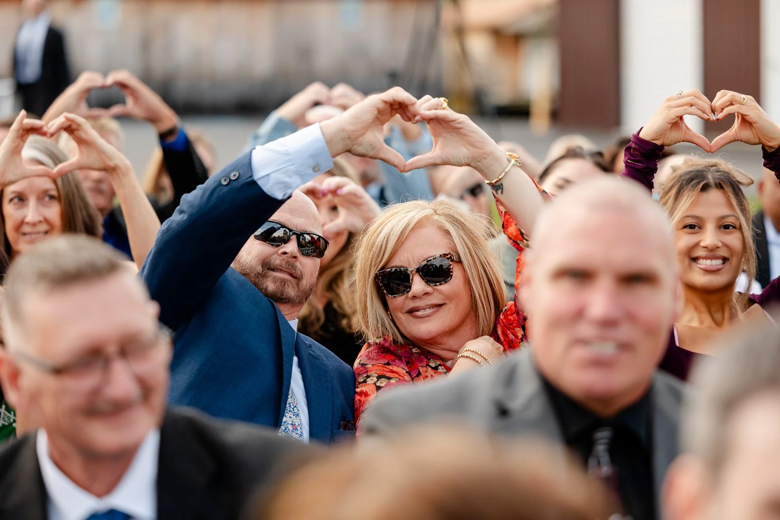 Group of people at an outdoor event making heart shapes with their hands above their heads, smiling and wearing formal and casual attire.