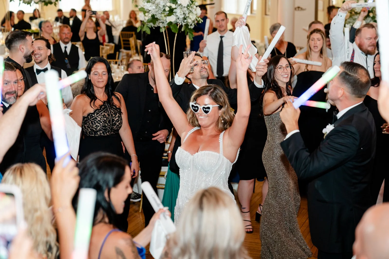 People dancing and celebrating at a wedding reception, some wearing sunglasses and holding glow sticks.