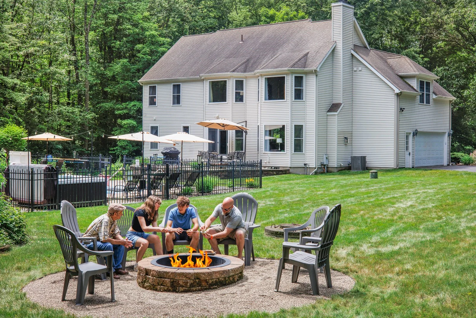 A family of four sitting around a fire pit in a green backyard, playing a game, with a white house, patio, and trees in the background.