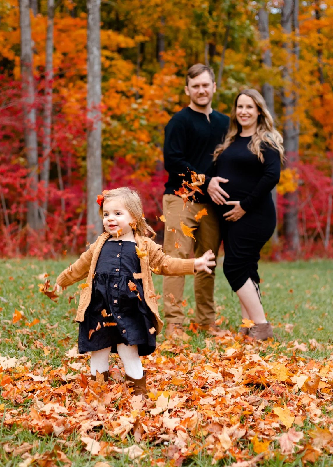 A family of three enjoying fall in a park with colorful autumn trees. A young girl in a navy dress and tan cardigan throws fallen leaves, while her parents, a man and a woman, stand behind her smiling. The mother is pregnant, and the scene shows a pl