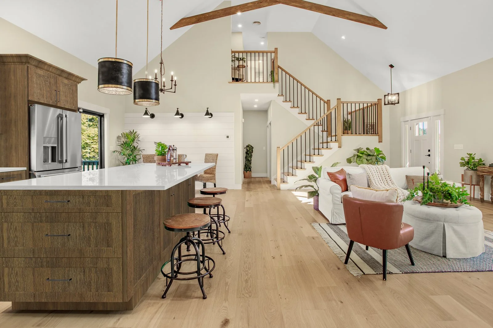 Open-concept living room and kitchen with wooden floors, staircase, white sofa, leather chair, kitchen island, and modern lighting fixtures.