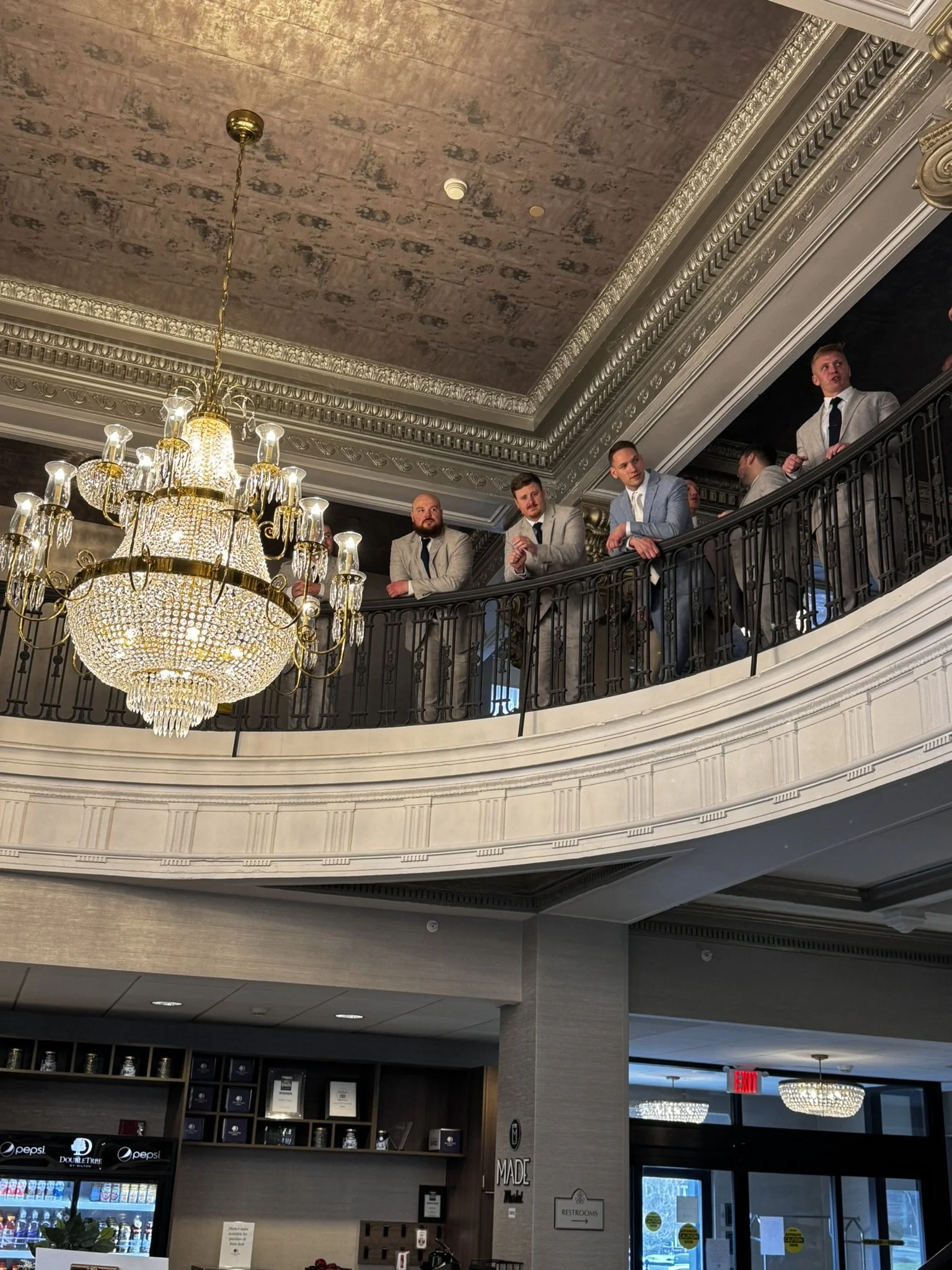 Men in suits standing on a balcony inside a building with ornate ceiling moldings and a large crystal chandelier.