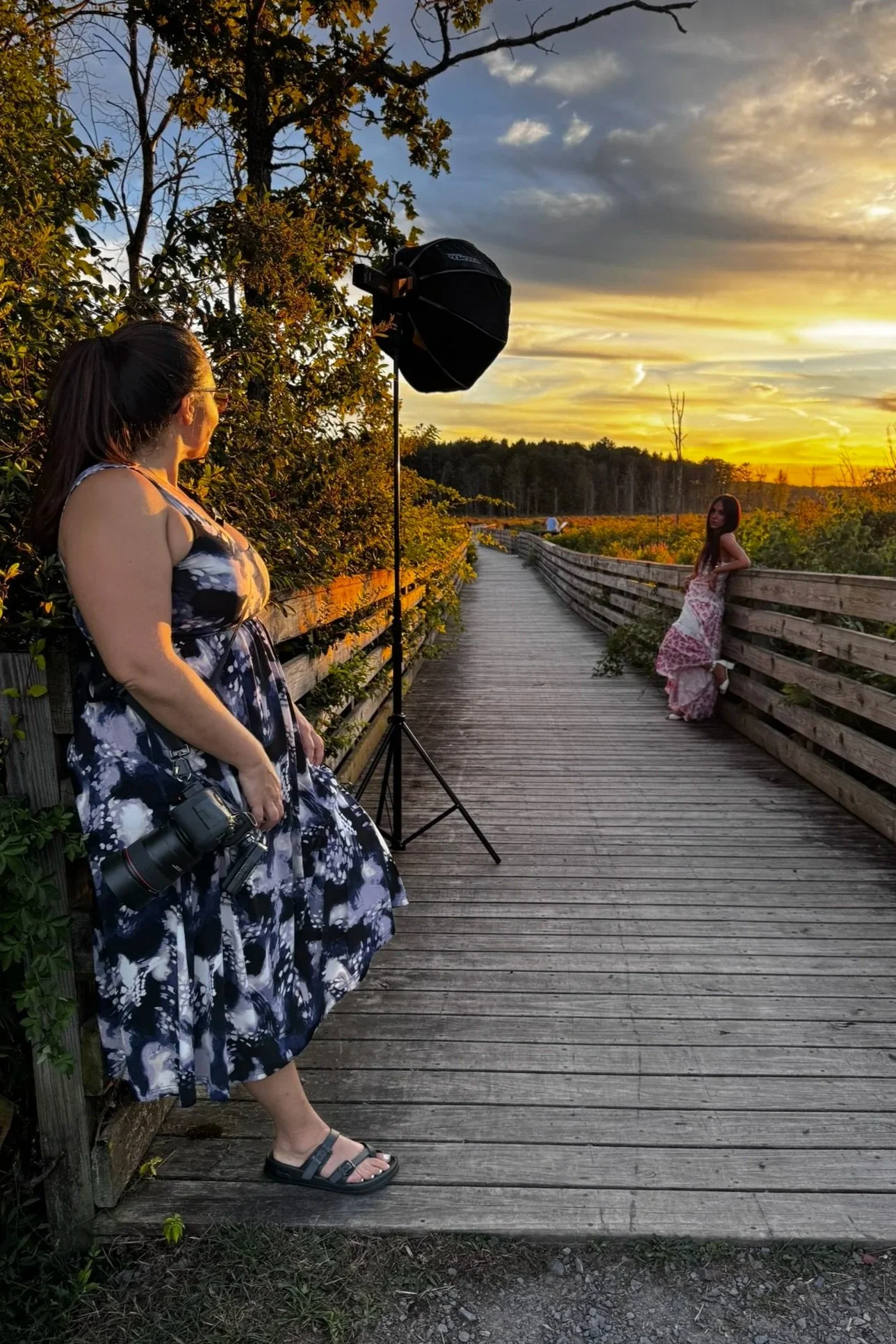 A woman with a camera around her neck takes photos of a woman in a pink and white dress leaning on a wooden railing during sunset on a wooden walkway surrounded by greenery.