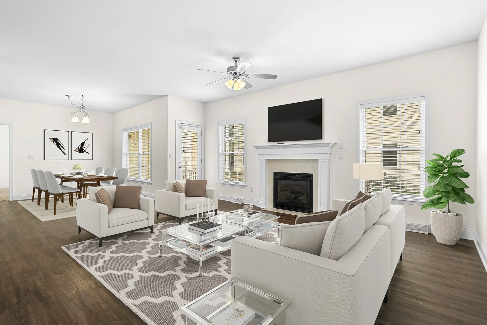 Bright living room with white walls, hardwood floors, white and beige furniture, a fireplace, wall-mounted TV, large windows with blinds, a patterned rug, and a large potted plant.