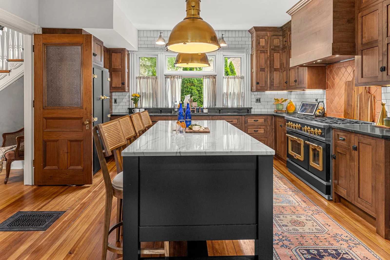 A kitchen with wooden cabinets, a black and white countertop, a black refrigerator, a large central island with a marble top, gold accents, a stove, and pendant lighting, with hardwood floors and a window with curtains letting in natural light.
