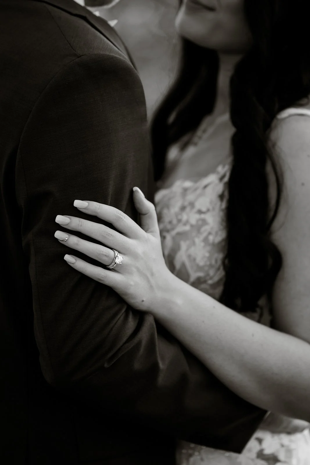 Close-up of a woman's hand resting on a man's arm, showing an engagement ring and wedding band.