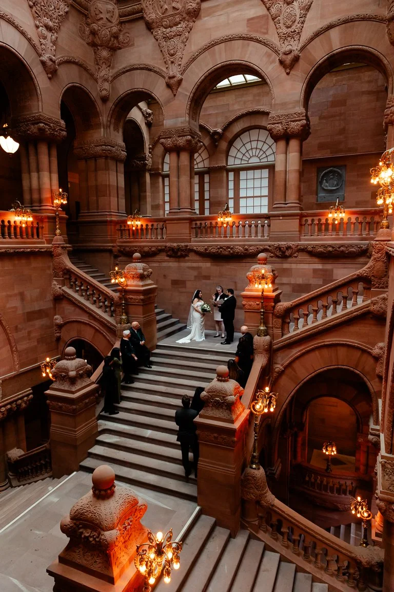 A wedding ceremony taking place on a grand staircase inside a historic building with ornate architecture and warm lighting.