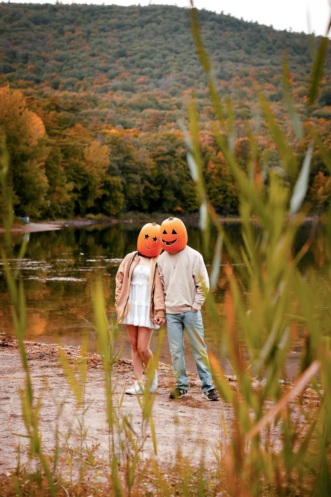 A couple stands by a lake with autumn-colored trees and mountains in the background. They are wearing pumpkin-shaped masks with carved faces, and holding hands.
