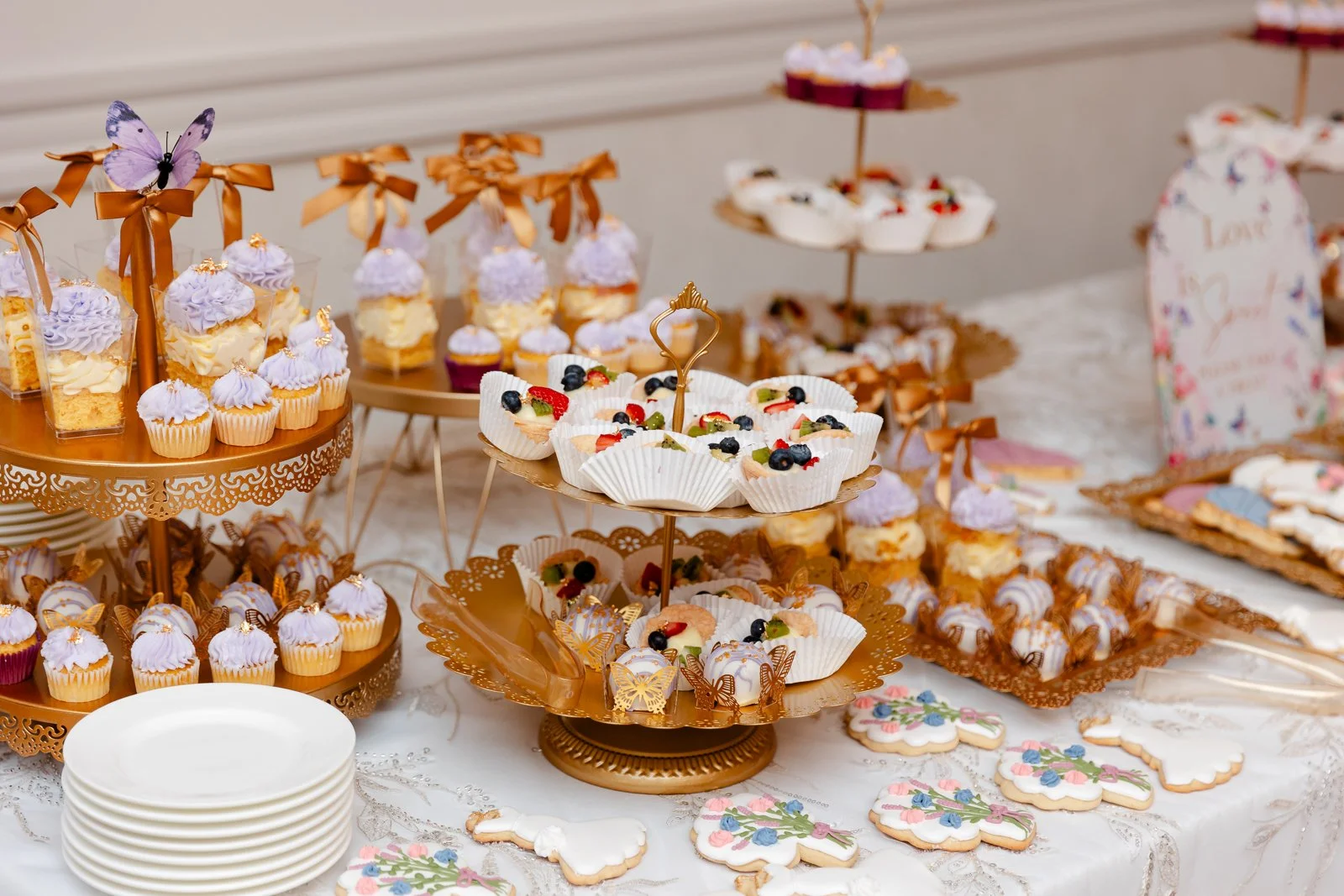 Desserts at a celebration table, including cupcakes with purple frosting, fruit-topped mini cheesecakes in white cupcake wrappers, cookies with floral icing, and layered desserts with ribbons and butterfly decorations.