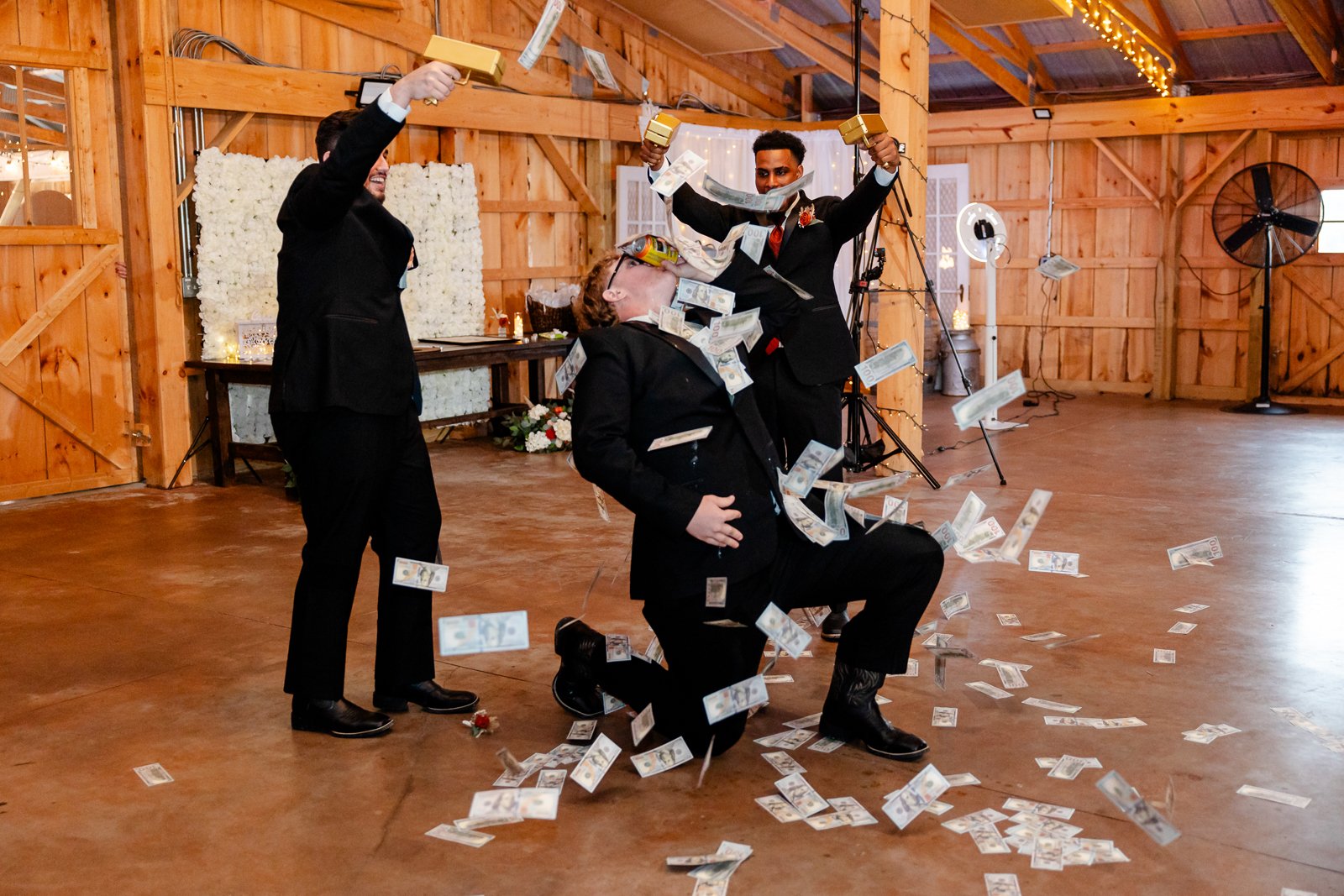 Three men dressed in suits celebrating with fake money at a rustic indoor event, with one kneeling, one standing, and one mid-air, all smiling.