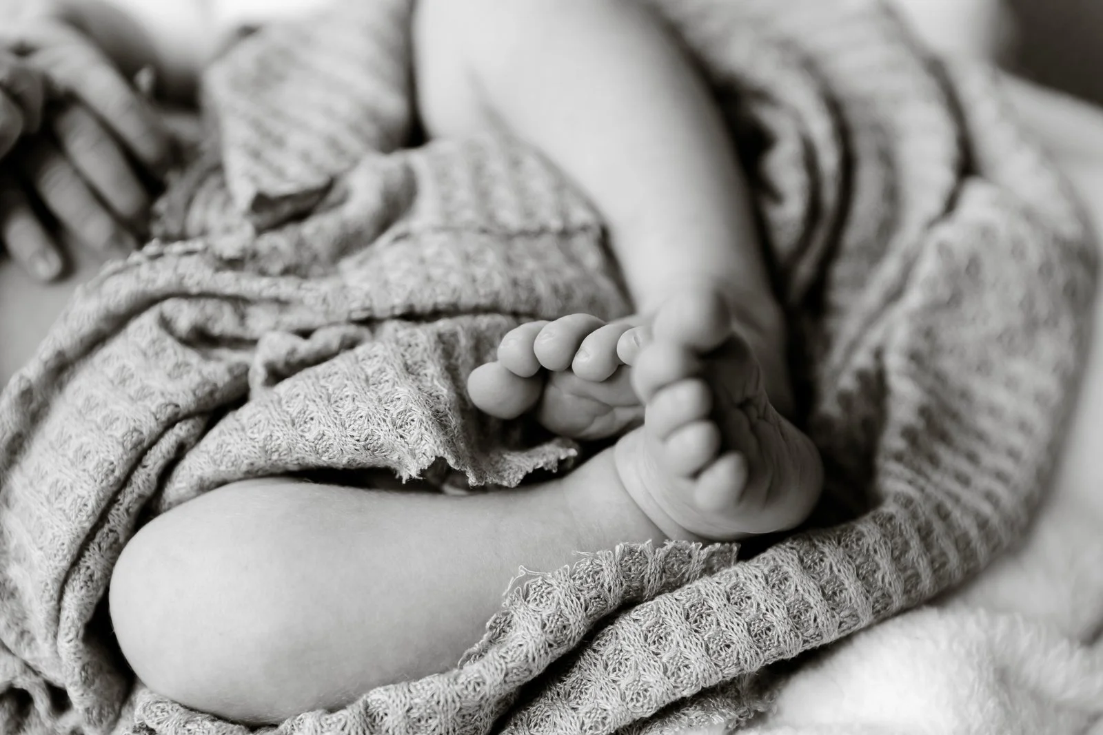 Black and white close-up photo of a baby, focusing on the baby's foot and curled toes, with the baby wrapped in textured blankets.