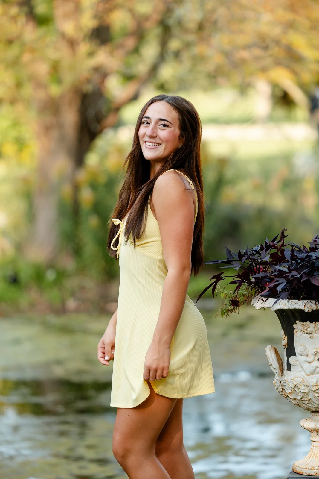 A young woman in a yellow dress standing outdoors near a pond, smiling at the camera with trees in the background.