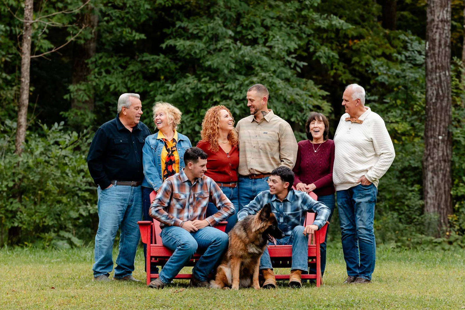 A multigenerational family gathered outdoors in a forested area, standing and sitting around a red bench with a German shepherd dog, all smiling and engaging with each other.