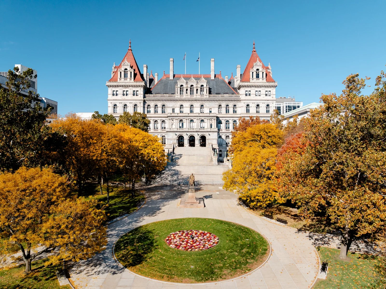 Photo of a large, historic, white stone government building with red pointed roofs, surrounded by trees with autumn-colored leaves, a statue, and a circular flower bed in the park in front of the building.