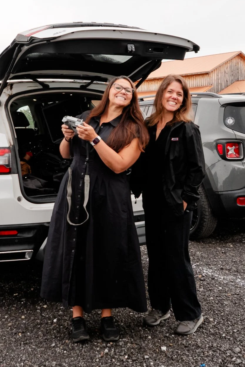 Two women smiling and posing with a drone near an open trunk of a white vehicle, with another gray vehicle and wooden building in the background.