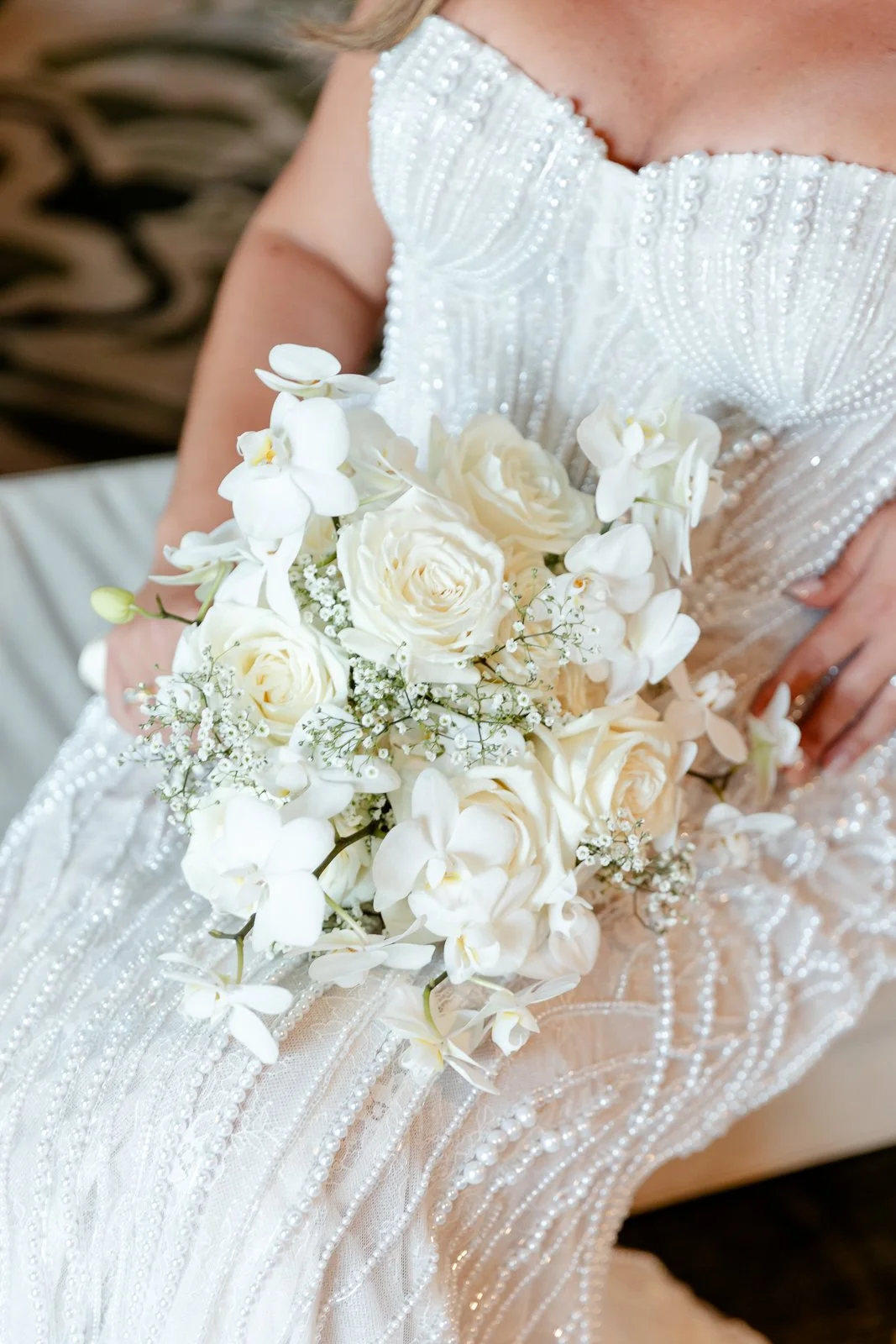 A bride holding a bouquet of white roses, orchids, and baby's breath, wearing a dress with pearl embellishments.