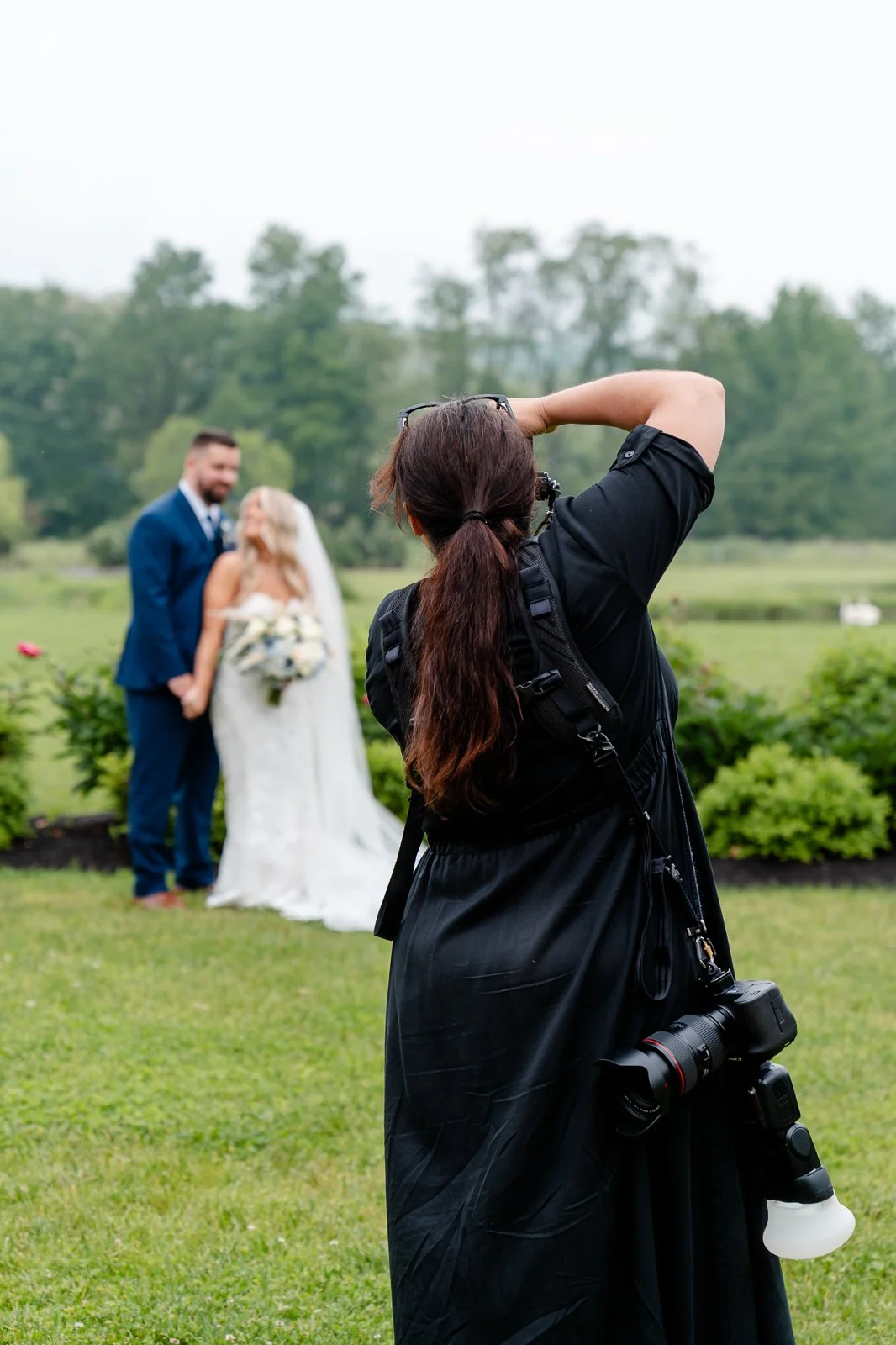 Photographer taking a picture of a bride and groom standing outdoors on a cloudy day with green trees and grass in the background.