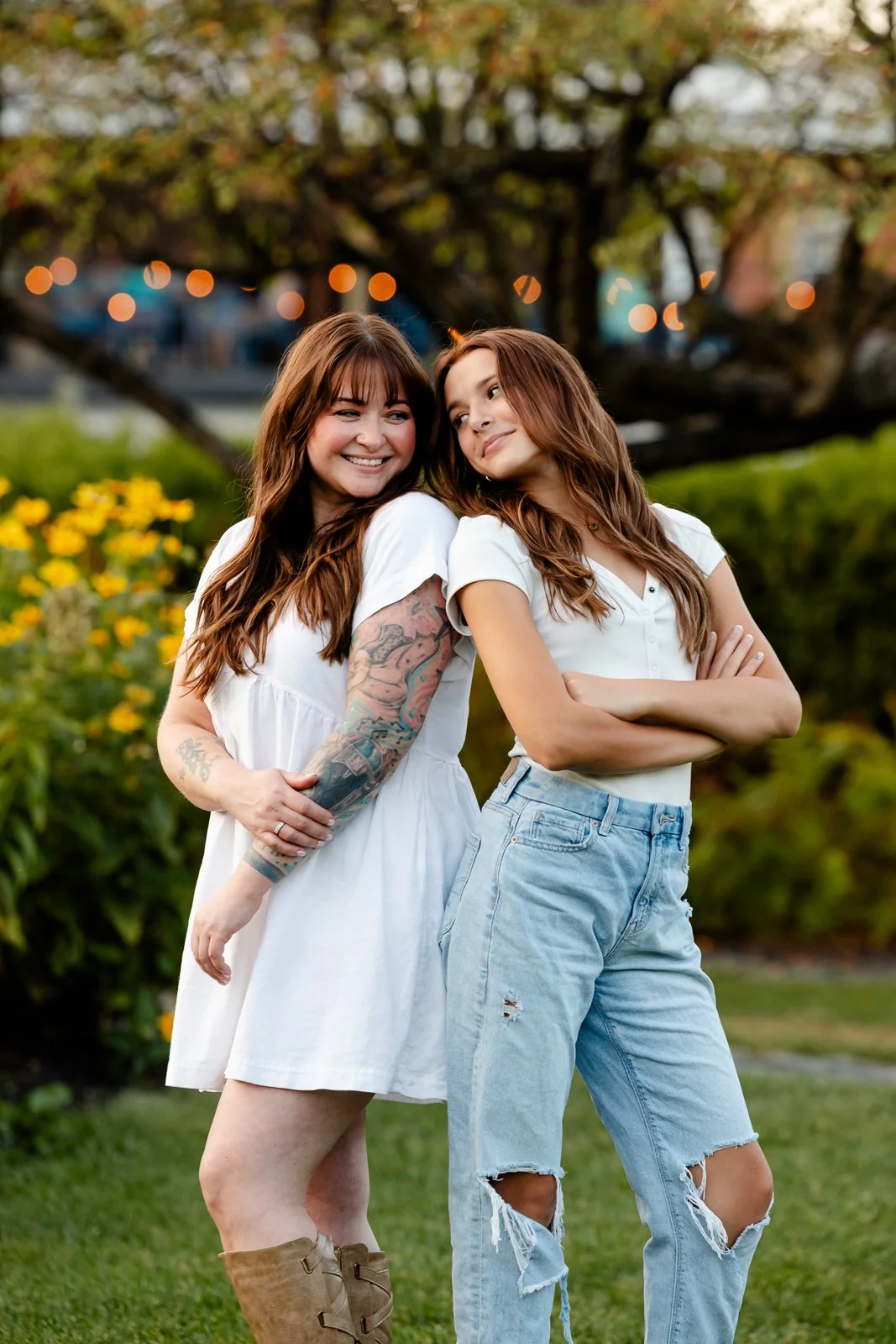 Two women, one with long brown hair and tattoos, smiling and standing close to each other in a garden with yellow flowers and a large tree in the background.