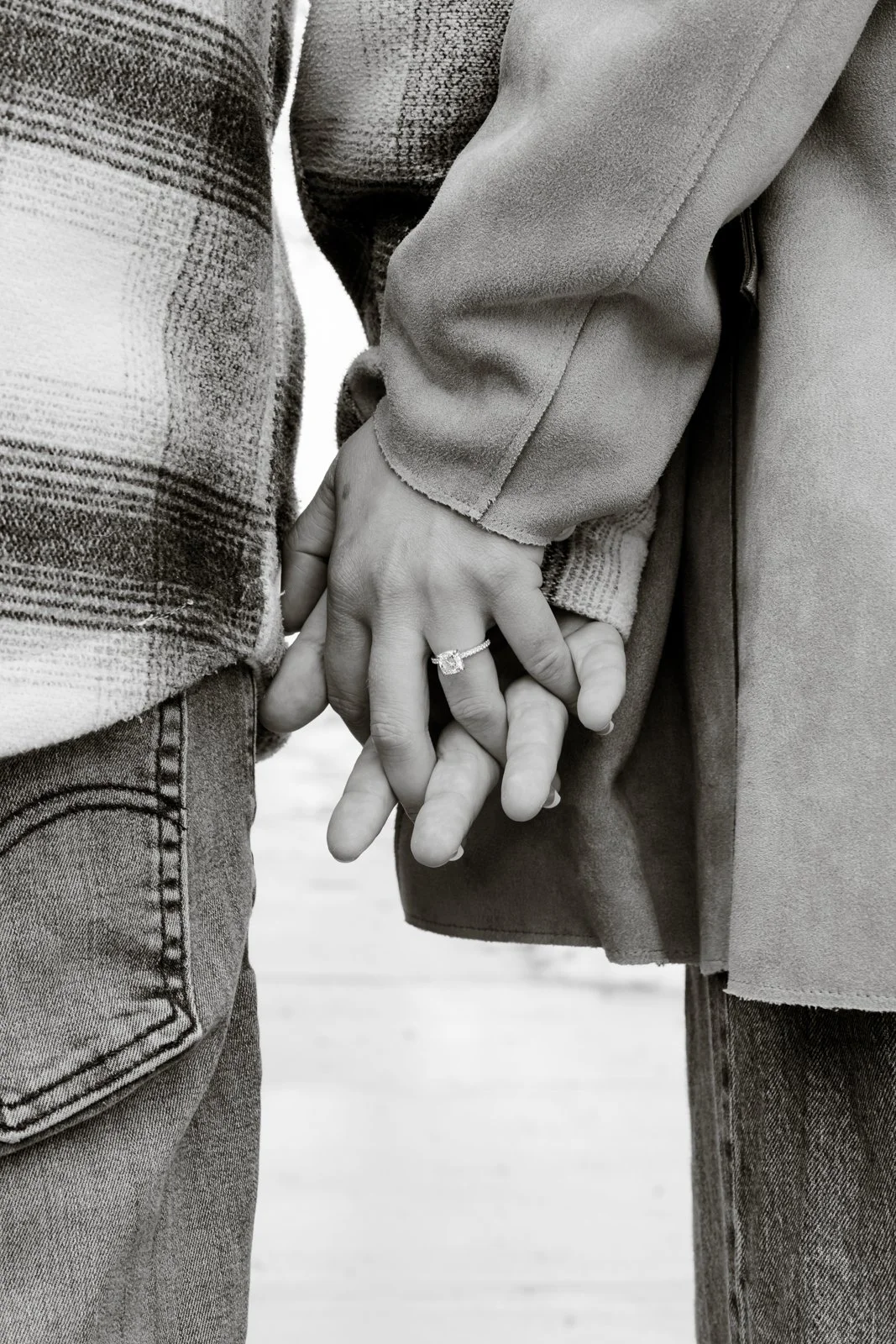 Close-up of a couple holding hands, with the woman wearing a ring. Both are dressed in casual outerwear and jeans.