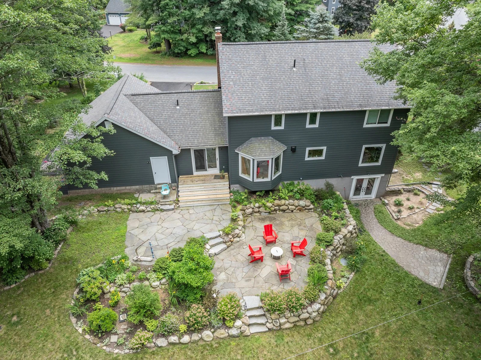 Aerial view of a suburban house with a backyard patio featuring four red Adirondack chairs arranged around a small table, surrounded by greenery and a curved stone pathway leading to the house.