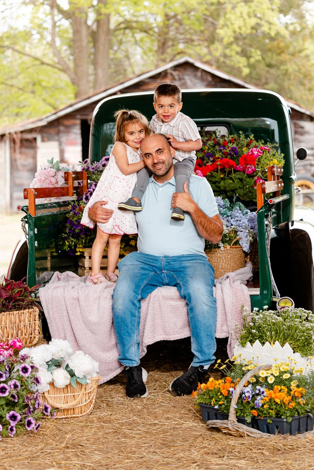 A man with two children in a vintage truck filled with colorful flowers during daytime in a rural setting.